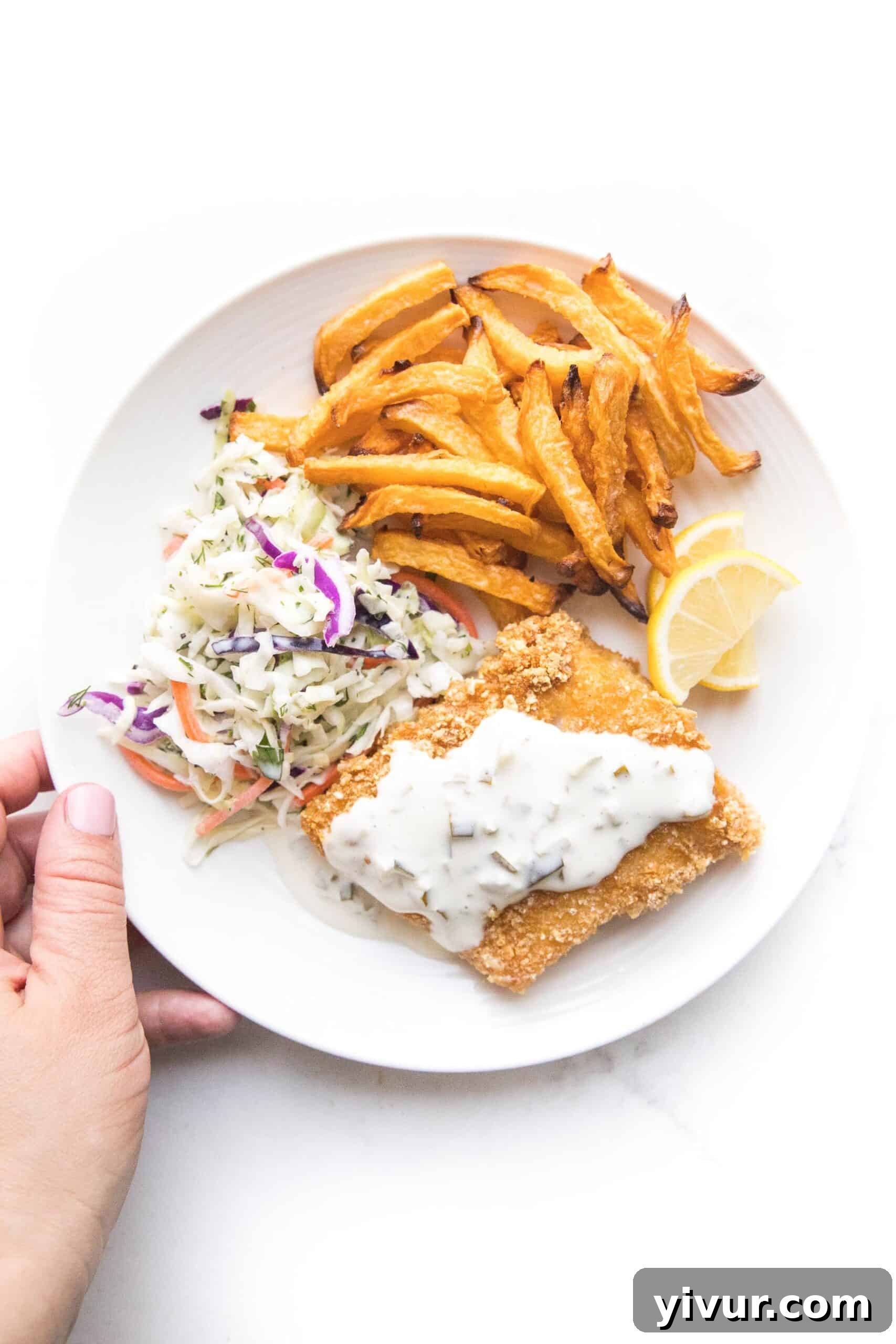 A hand holding a plate with a golden brown keto fish filet with keto fries, coleslaw, lemon wedges, and tartar sauce on top ready to serve, symbolizing a complete meal.