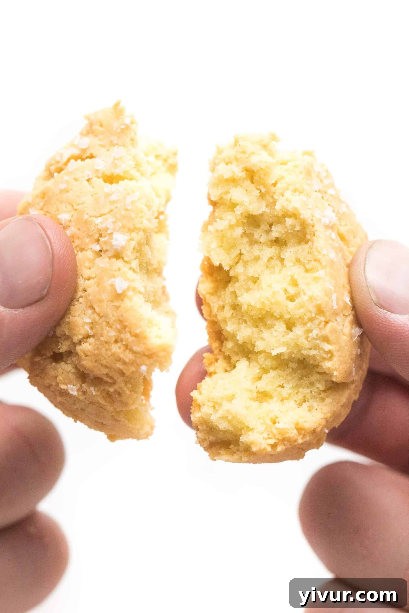 Hand breaking apart a golden-brown almond flour keto biscuit, revealing its flaky texture on a white background.