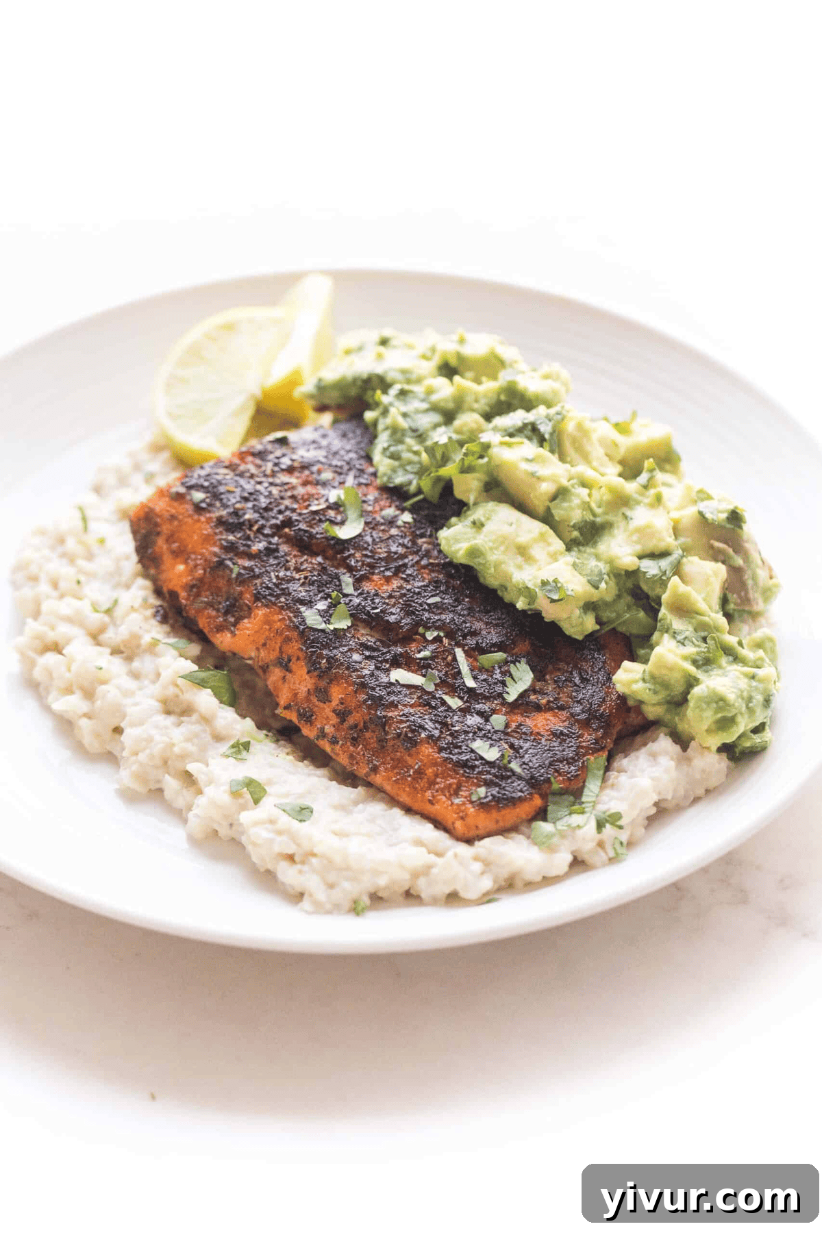 A white plate with a serving of cauliflower rice, a blackened salmon fillet, and a dollop of chunky guacamole, garnished with lemon slices.