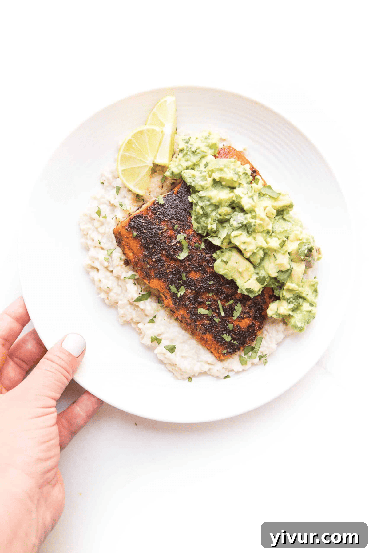 A hand holding a white plate with cauliflower rice, a blackened salmon fillet, and chunky guacamole, garnished with lemon slices.