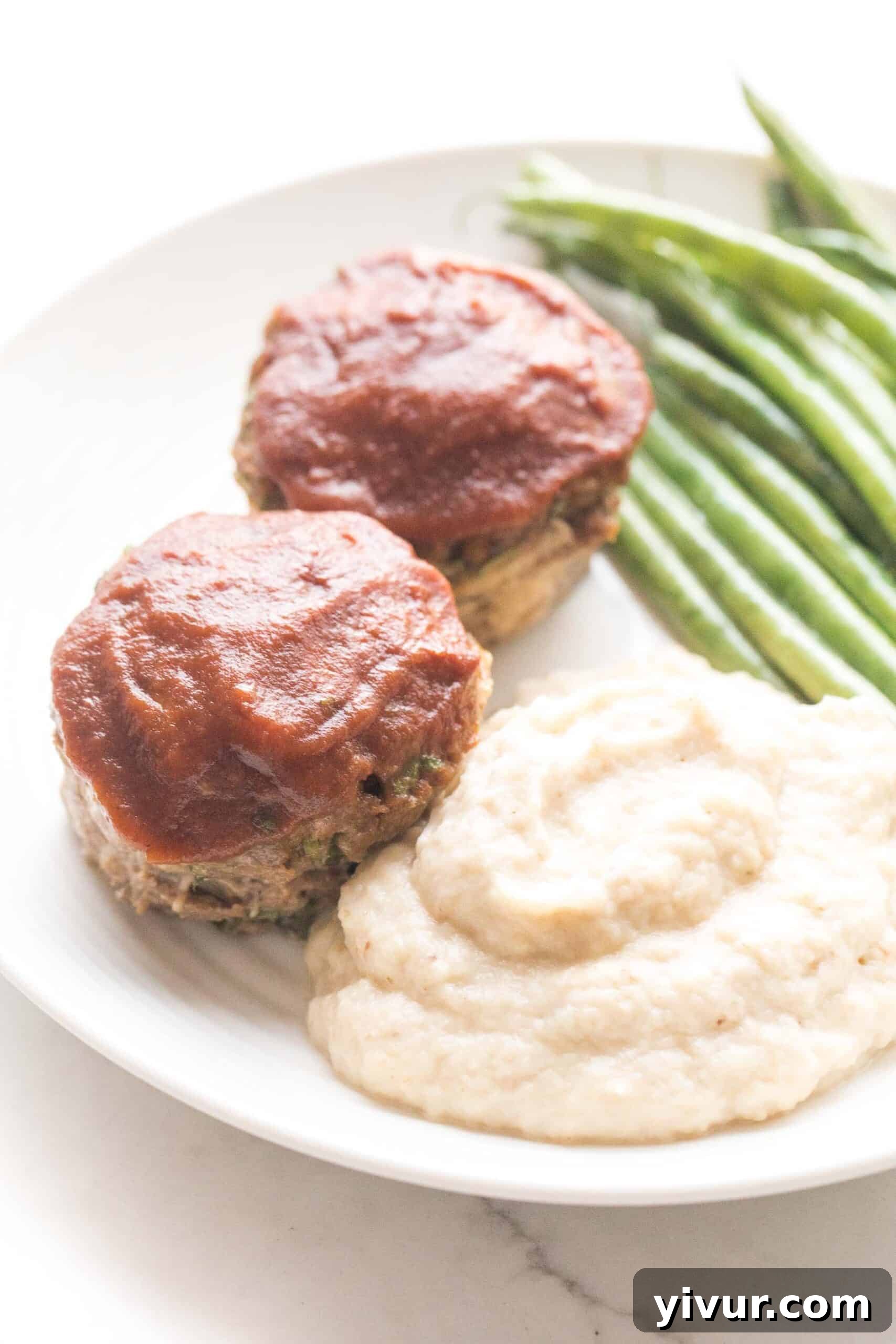 Quick Keto Meatloaf Muffin Bites 2 Mini meatloaf muffins topped with sugar-free ketchup, served with roasted green beans and creamy mashed cauliflower on a white plate, showcasing a healthy and delicious keto meal.
