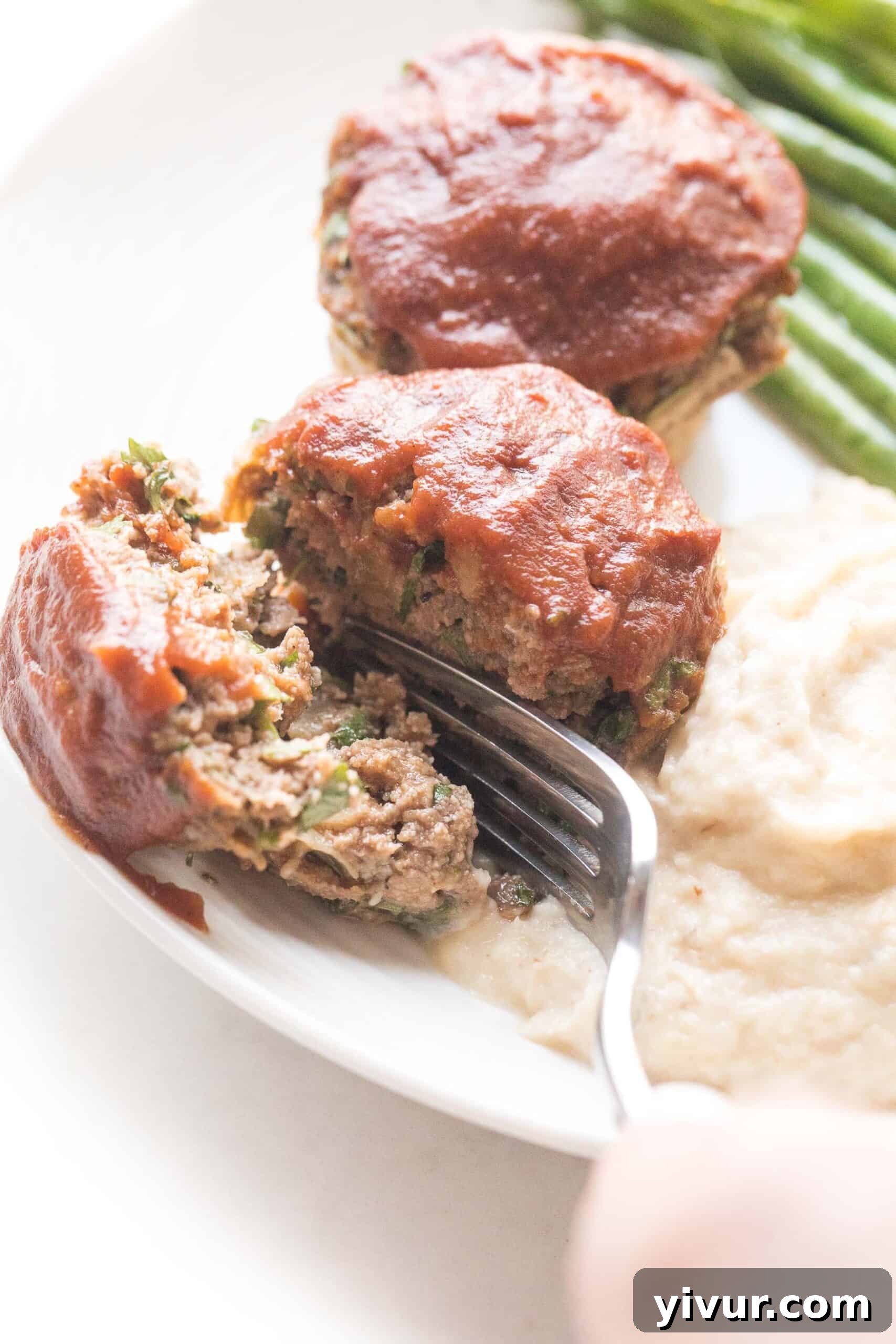 Quick Keto Meatloaf Muffin Bites 5 A knife slicing into a mini meatloaf muffin topped with ketchup, served with roasted green beans and mashed cauliflower on a white plate, highlighting its tender texture.