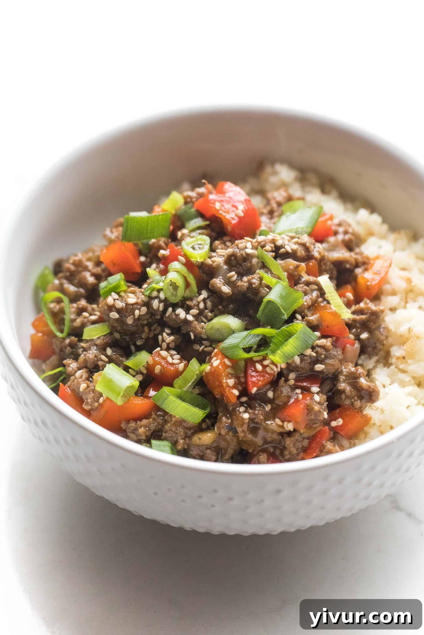 Umami-Rich Whole30 Keto Asian Beef Bowls 2 A beautifully presented bowl of ground Asian beef and bell peppers, generously topped with vibrant green onions, served over a bed of fluffy cauliflower rice. The white bowl highlights the rich colors of the meal.