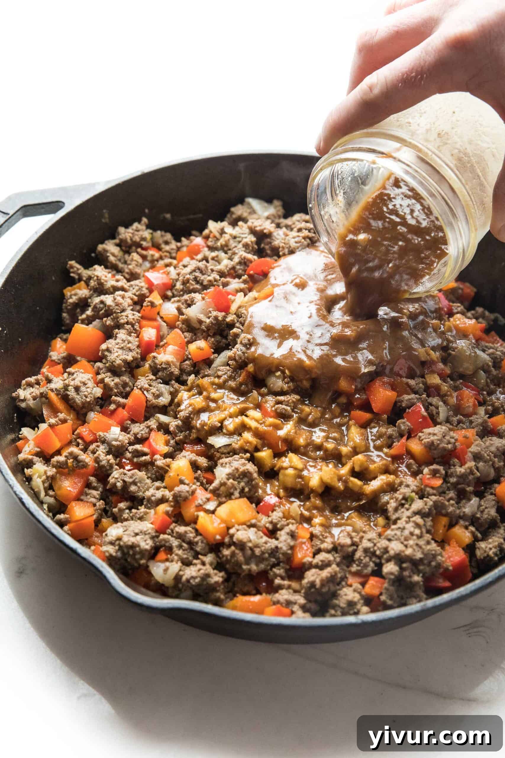 Umami-Rich Whole30 Keto Asian Beef Bowls 3 A close-up shot capturing a rich, homemade stir-fry sauce being poured over browned ground beef and colorful bell peppers simmering in a rustic cast iron skillet, promising deep flavor.