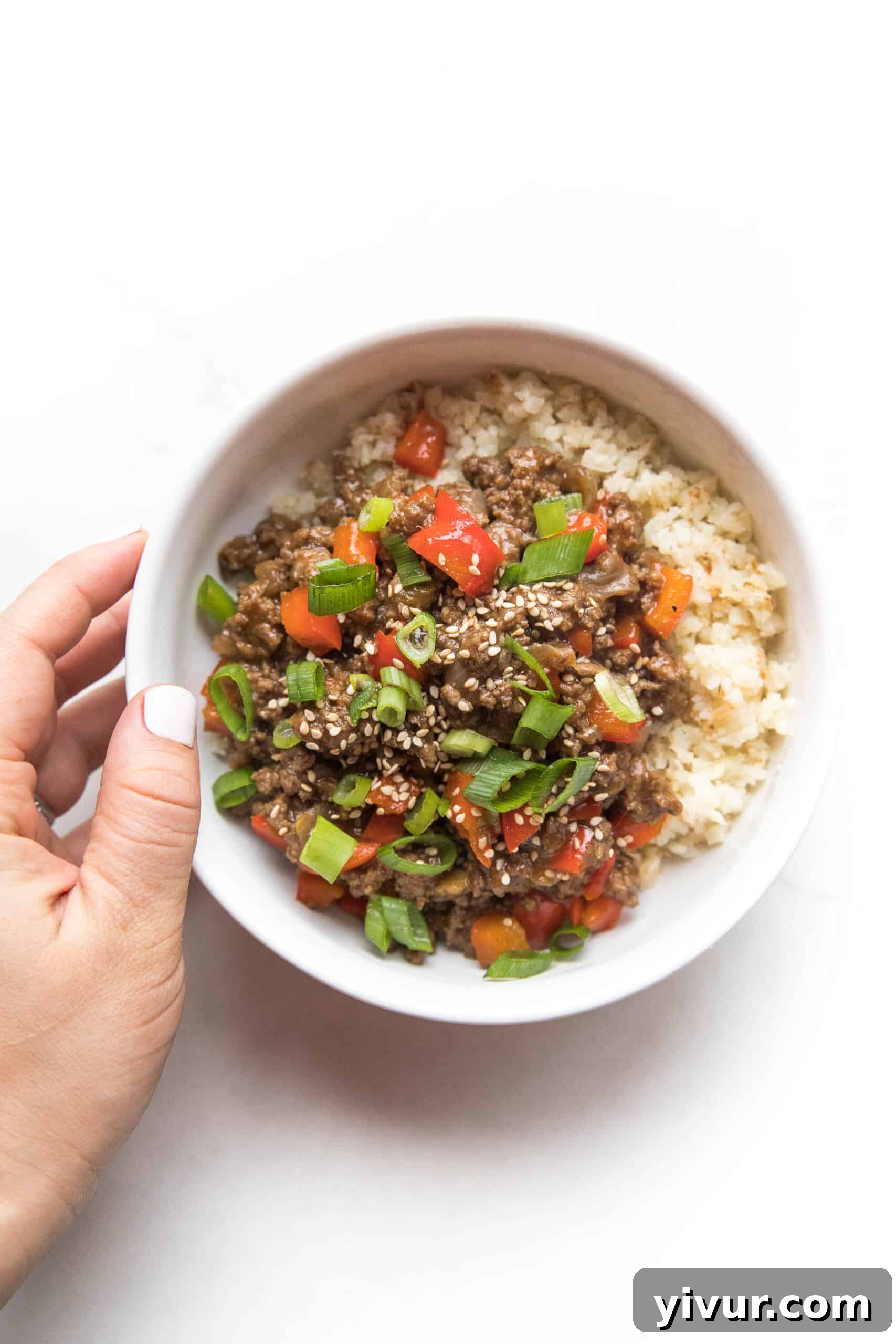 Umami-Rich Whole30 Keto Asian Beef Bowls 4 A hand gently holds a white bowl filled with ground Asian beef, bell peppers, and cauliflower rice, beautifully garnished with fresh green onions. The focus is on the delicious texture and inviting presentation.