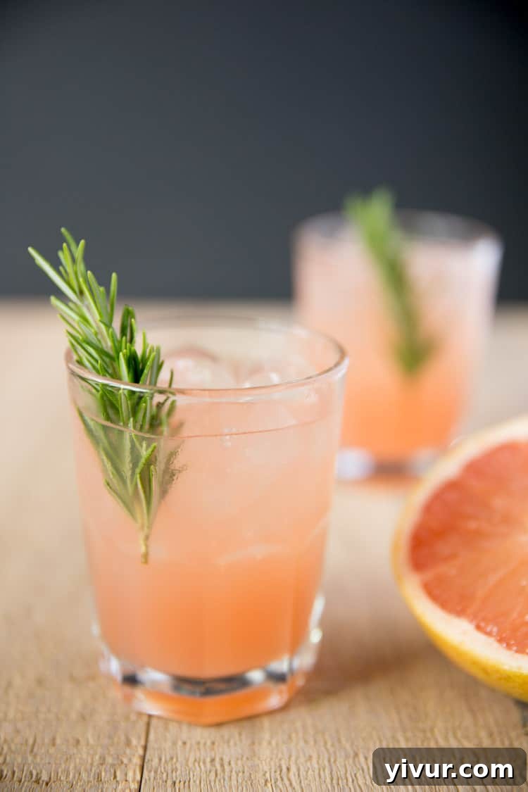 Two glasses of vibrant rosemary greyhound cocktails, garnished with fresh rosemary sprigs, sit alongside a perfectly halved ruby red grapefruit.