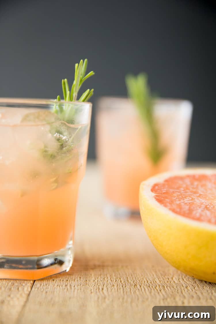 Two glasses of finished rosemary greyhound cocktails, elegantly garnished with rosemary sprigs, sitting beside a half grapefruit.