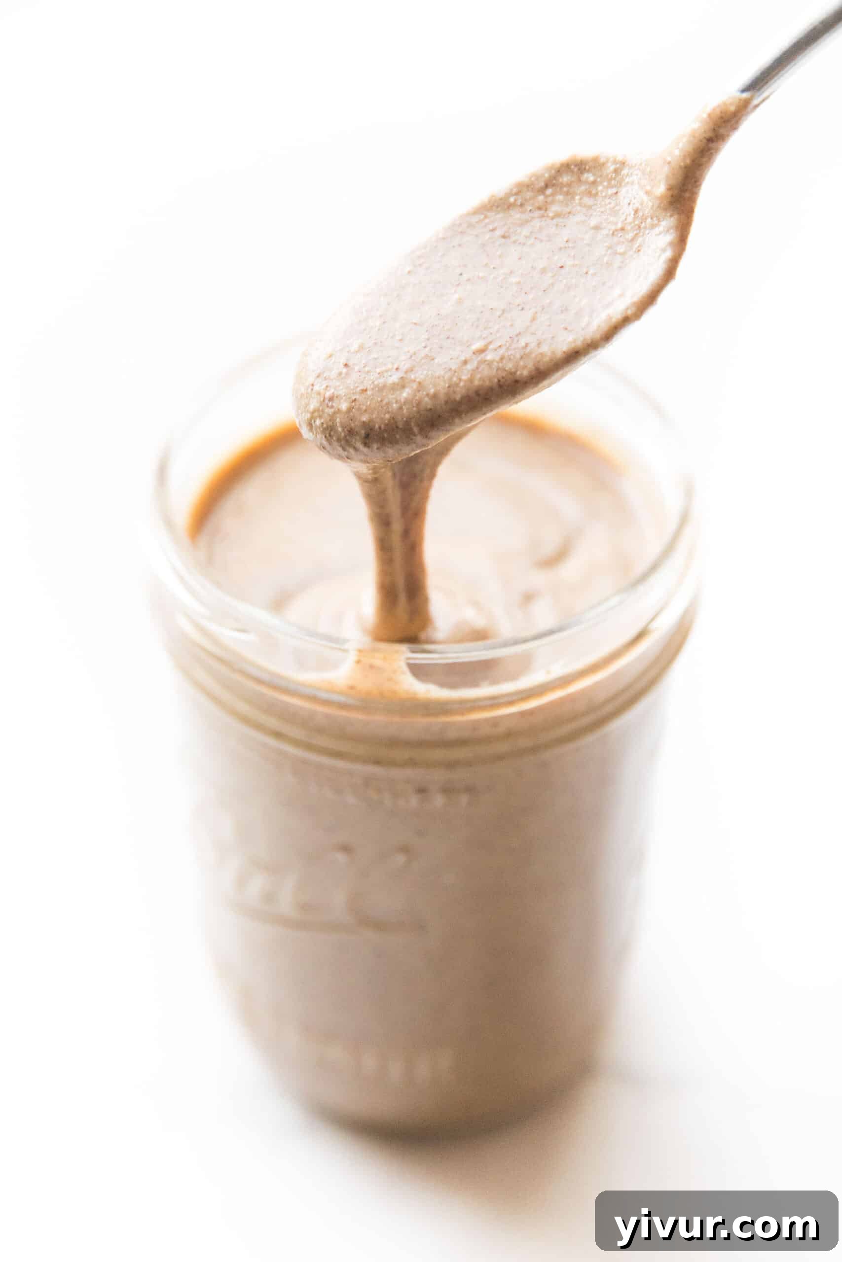 Spoon drizzling creamy mixed nut butter into a clear mason jar, showcasing its smooth texture and rich color.