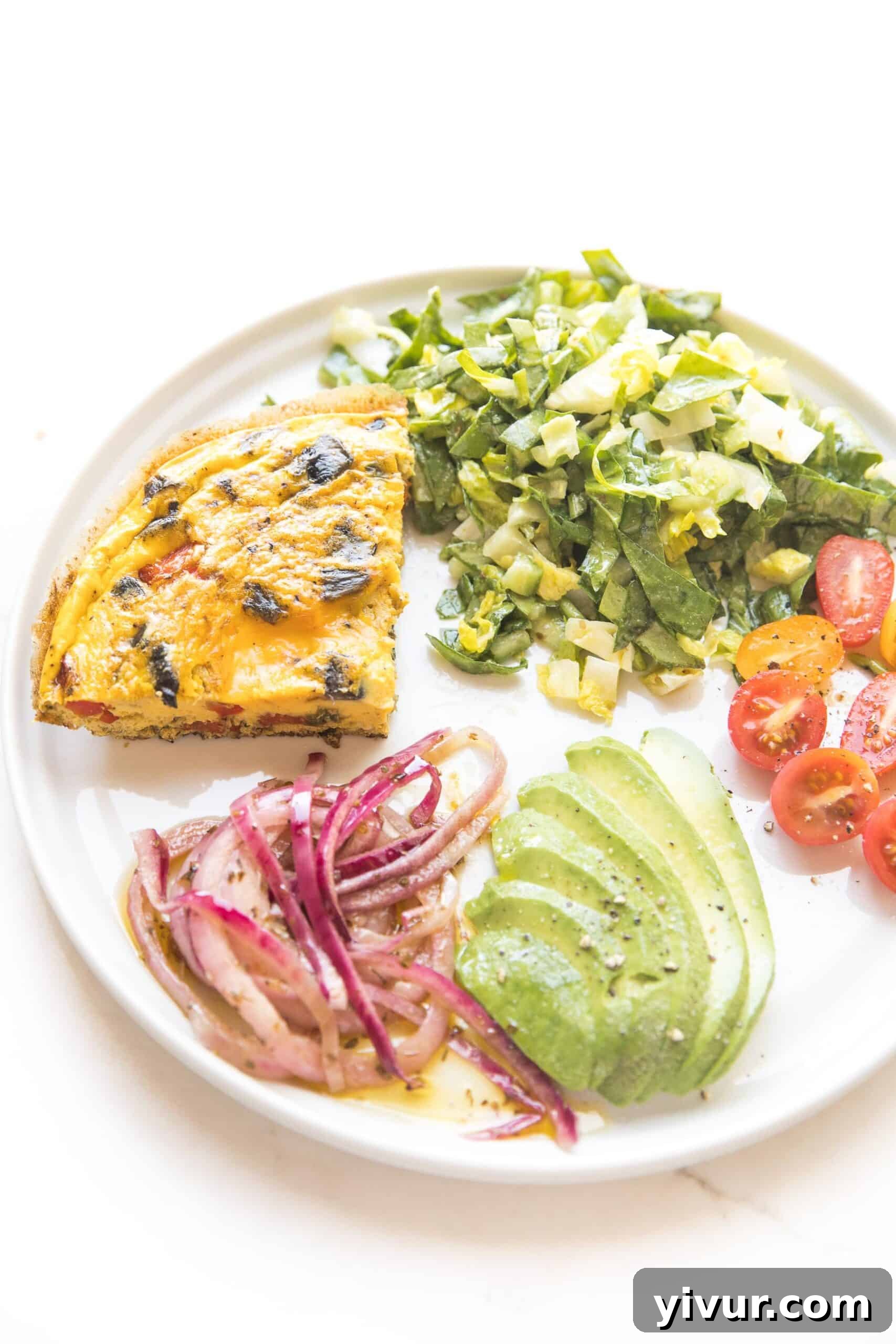 A slice of keto frittata on a pristine white plate, accompanied by a side of marinated red onions, creamy avocado slices, fresh tomatoes, and a crisp green salad, highlighting a complete meal.