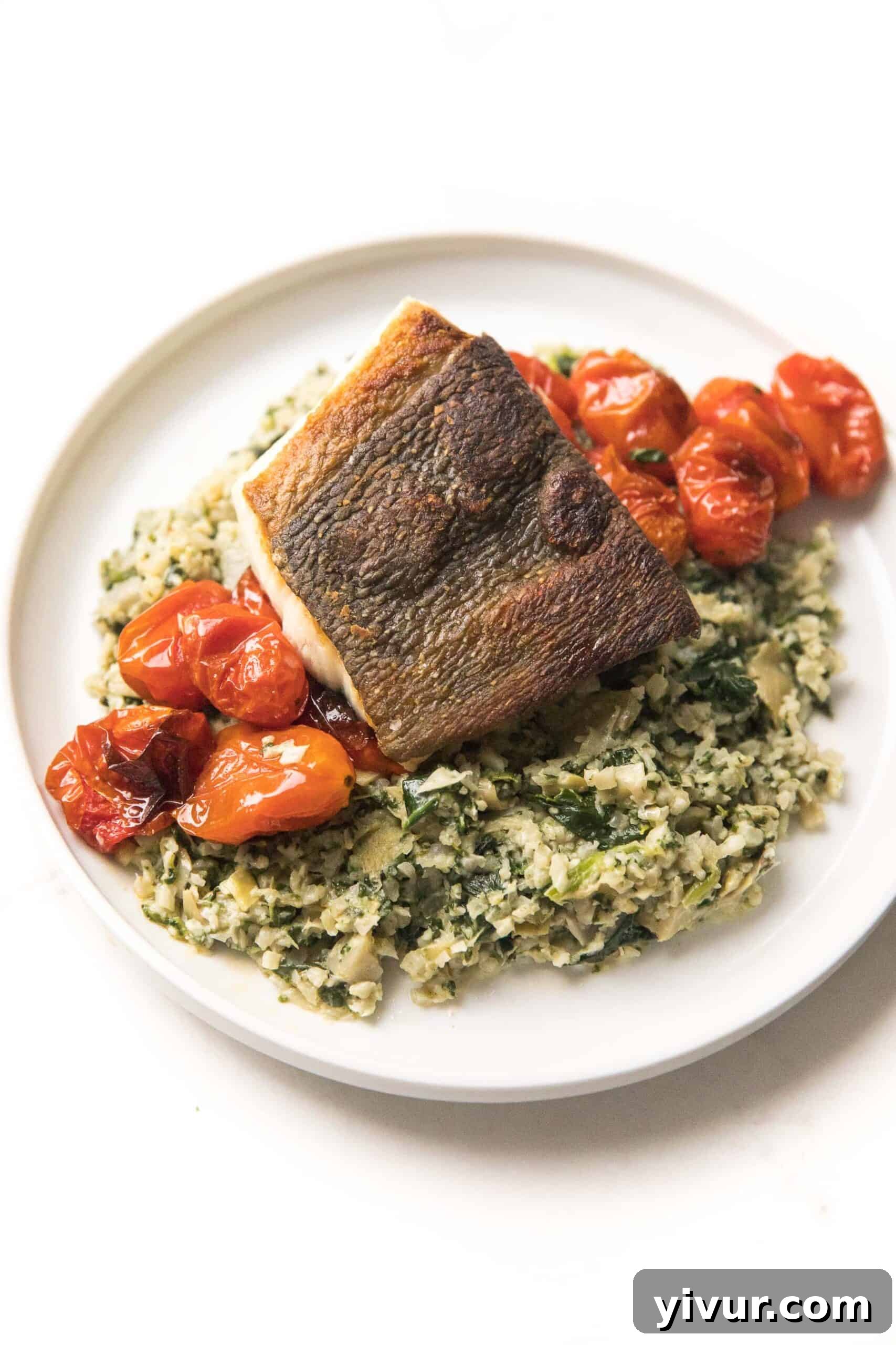Flaky Whole30 + Keto Halibut served on a bed of creamy Spinach Artichoke Cauliflower Rice Risotto, garnished with vibrant blistered tomatoes, presented on a pristine white plate.