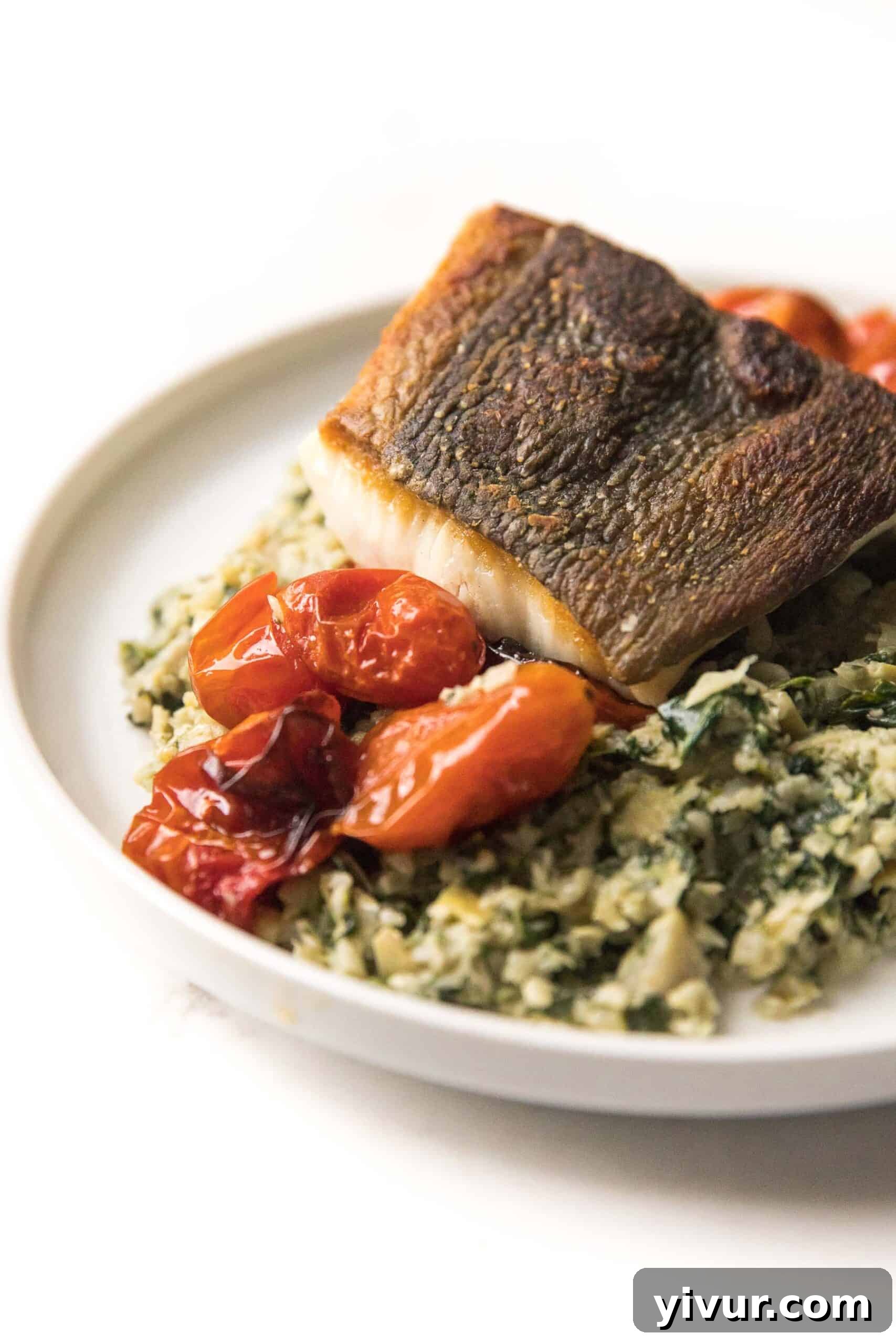 Close-up of Whole30 + Keto Halibut, perfectly seared with crispy skin, resting on creamy Spinach Artichoke Cauliflower Rice Risotto, accompanied by sweet blistered tomatoes.