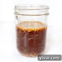 A wide-mouth glass jar filled with a dark brown stir fry sauce, featuring small pieces of chopped garlic and ginger floating in it.