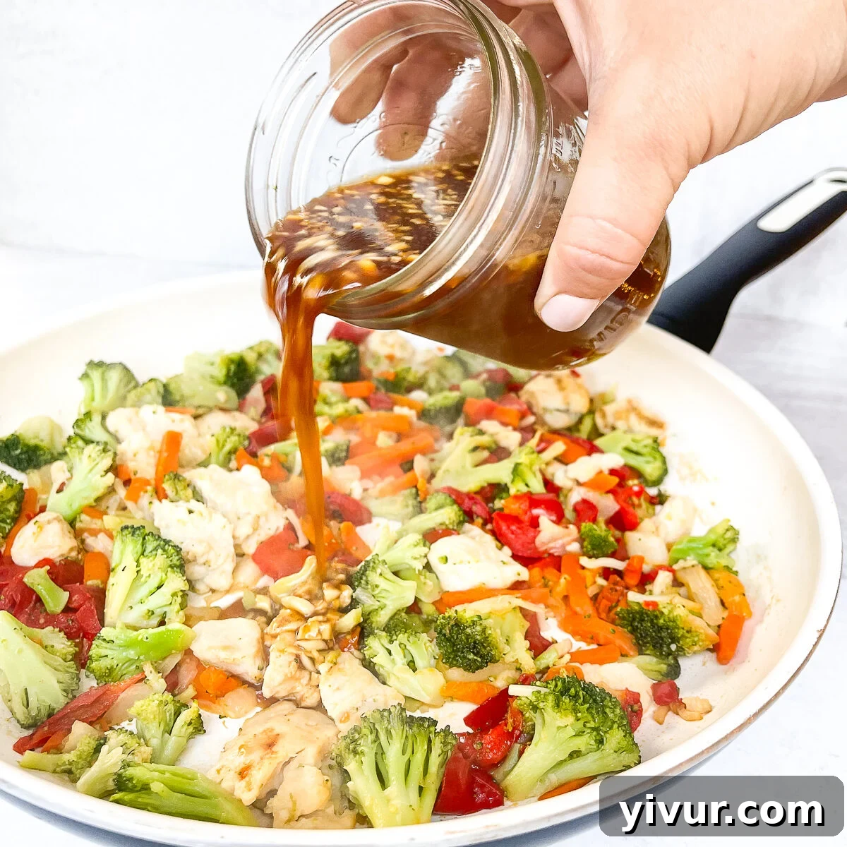 Thick stir fry sauce being poured over cooked chicken and vegetables in a white skillet.