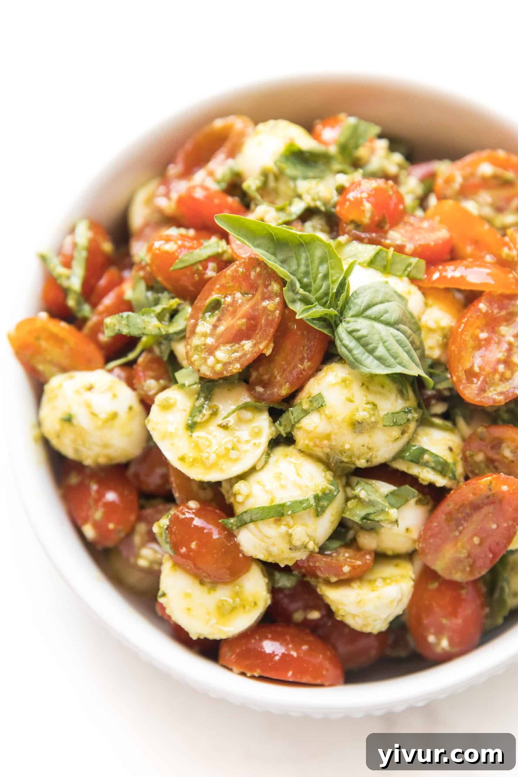 Brightly colored Keto Pesto Caprese Salad in a white serving bowl, showcasing cherry tomatoes, mozzarella balls, and fresh basil leaves.