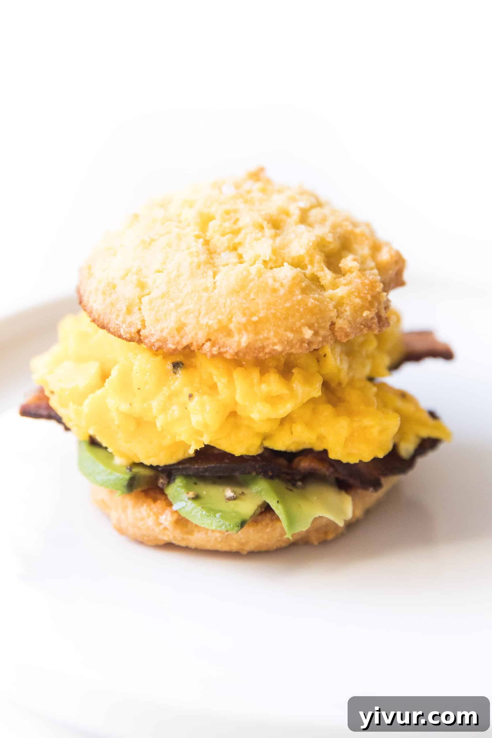 A perfectly assembled keto biscuit breakfast sandwich, featuring a golden biscuit, fluffy scrambled egg, crispy bacon, and creamy avocado slices, presented on a pristine white plate against a clean white background. It's a visually appealing and wholesome start to any day.