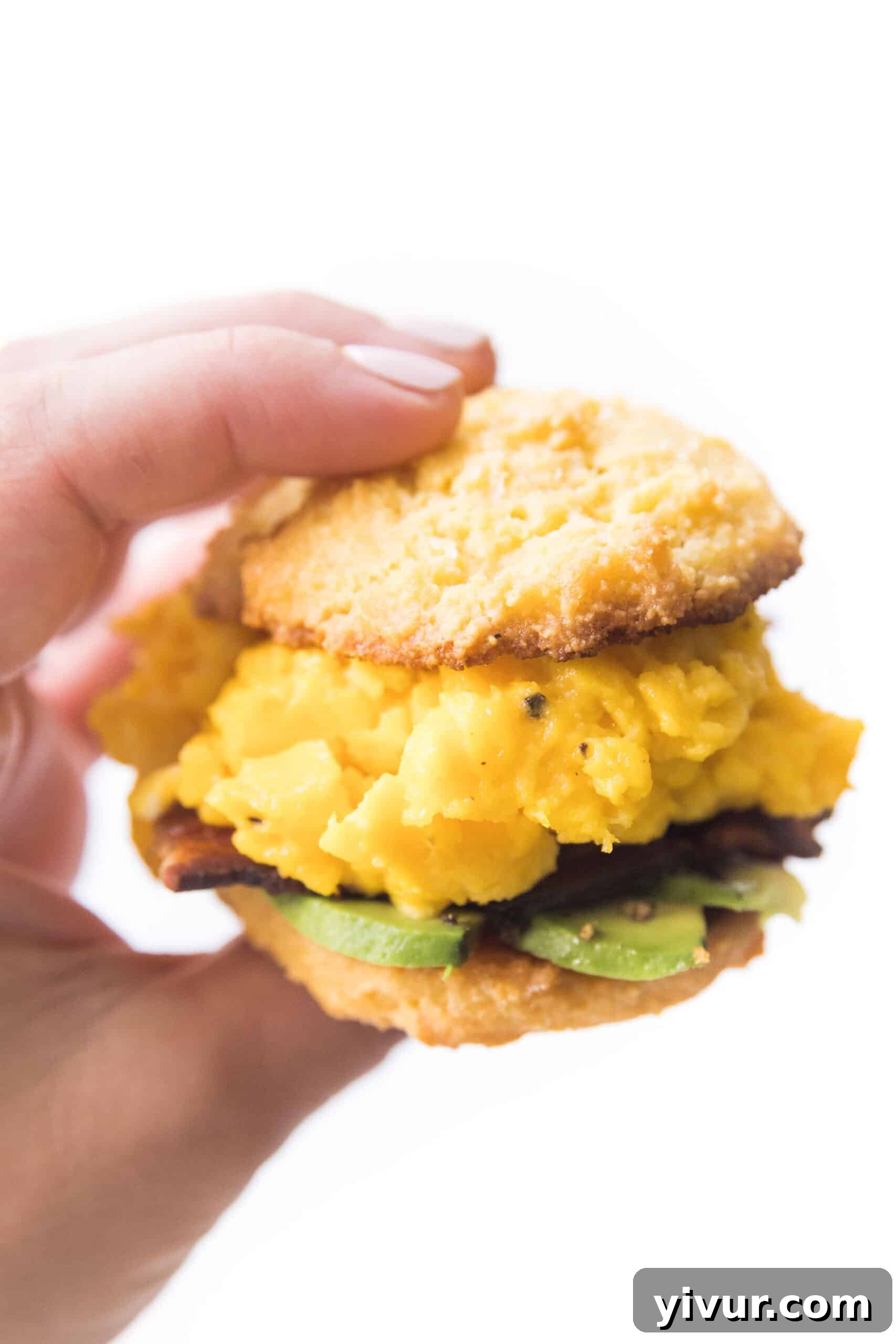 A close-up shot of a hand holding a delicious keto biscuit breakfast sandwich, showcasing the layers of egg, bacon, and avocado peeking out from between the halves of the golden-brown biscuit. The focus emphasizes the inviting texture and richness of the sandwich.