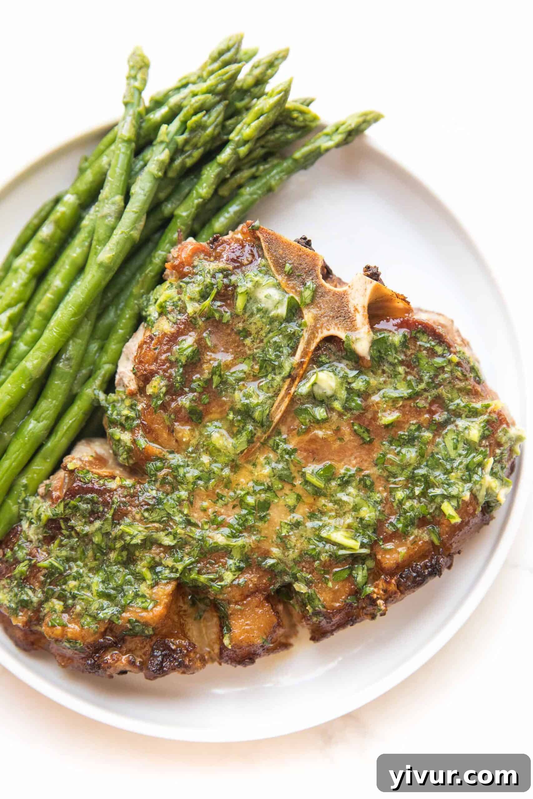 Juicy bone-in pork chop covered in melted lemon herb butter, garnished with fresh parsley and served with roasted asparagus on a white plate