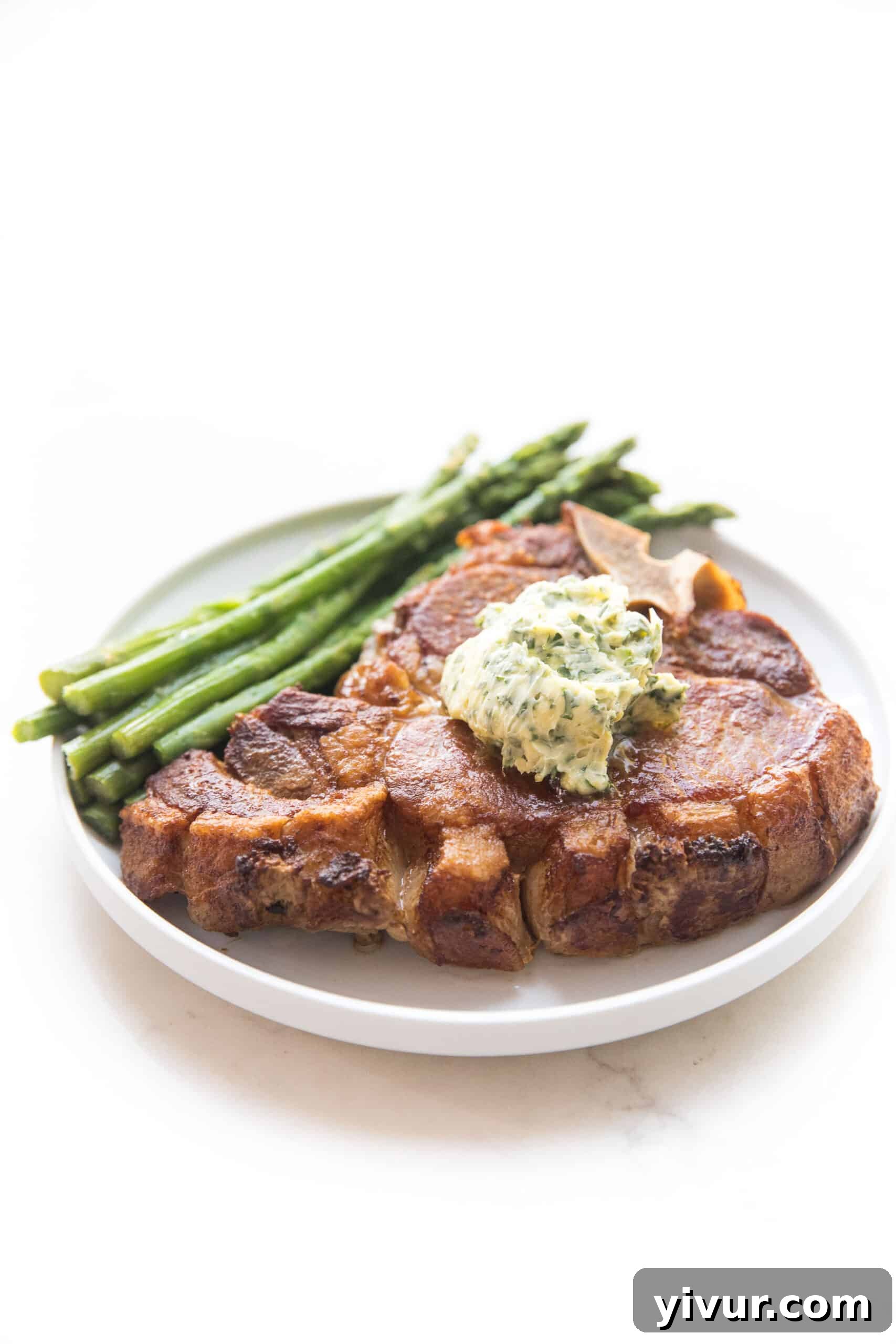 Cooked bone-in pork chop topped with a dollop of vibrant herb butter, accompanied by green asparagus on a clean white plate