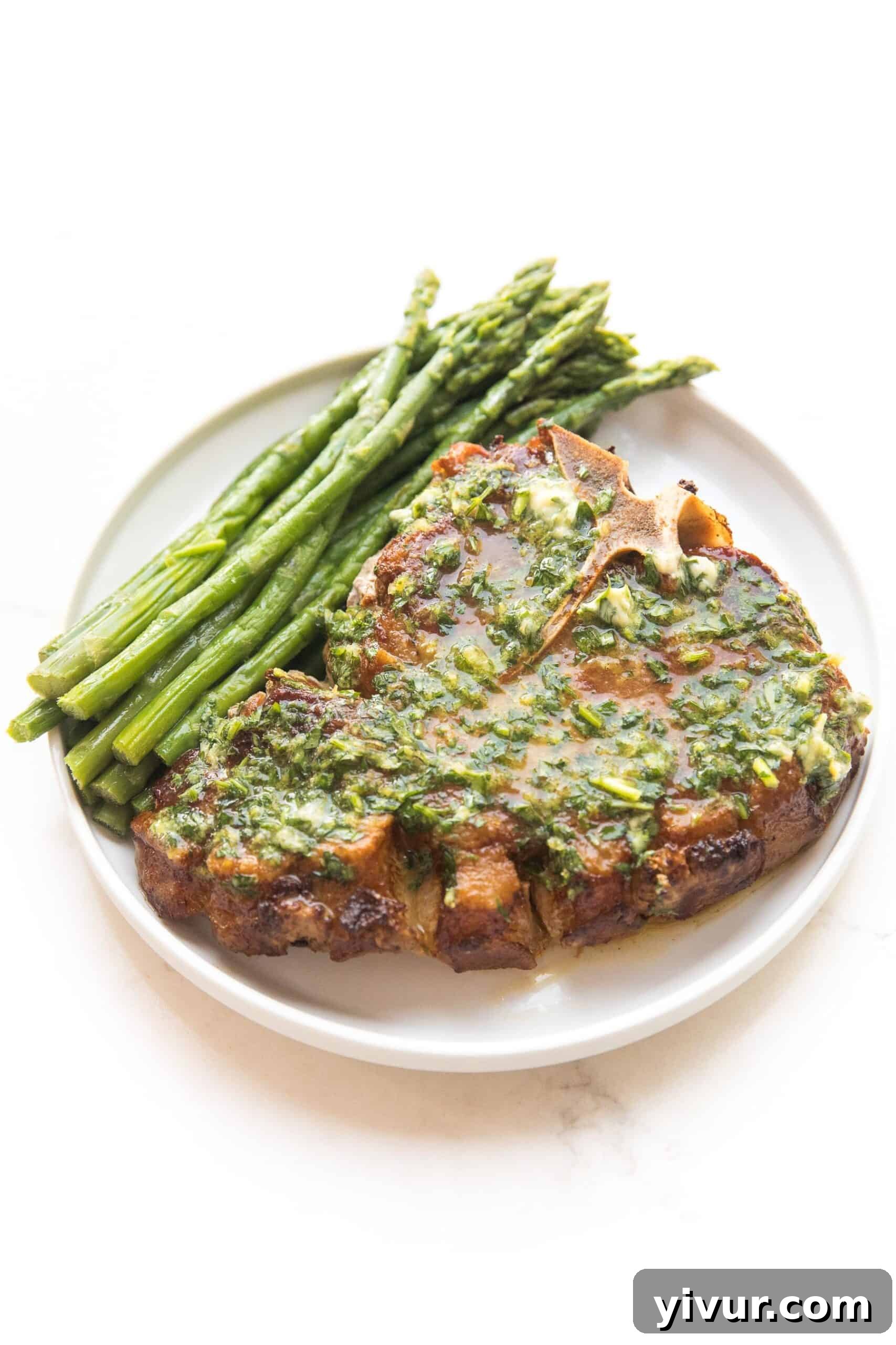 Three perfectly seared bone-in pork chops resting on a white plate, ready to be topped with lemon herb butter, alongside fresh asparagus