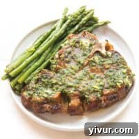 close up of a juicy pork chop covered in melted herb butter with parsley and asparagus on a white plate