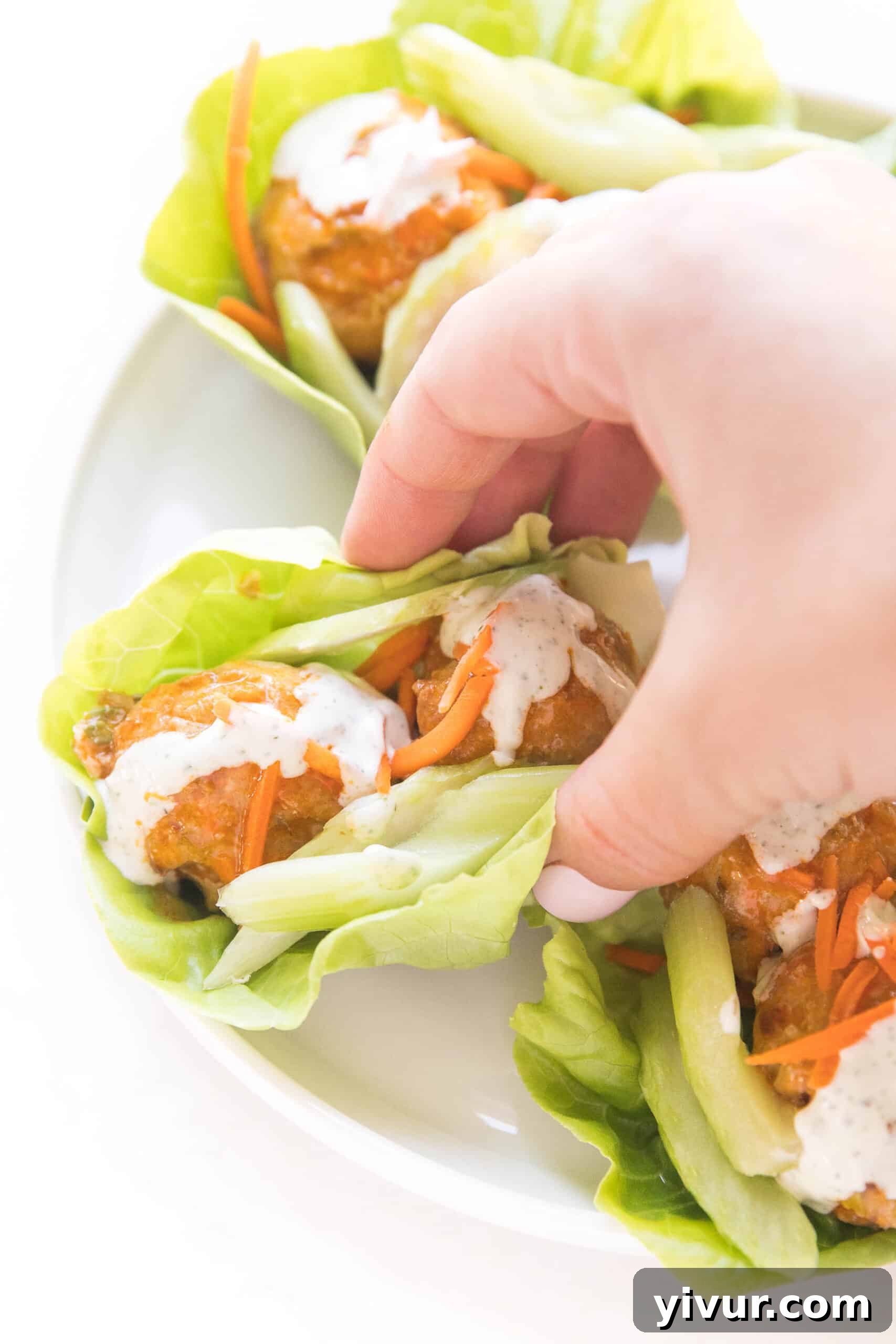 hand grabbing picking up keto buffalo turkey meatballs on lettuce wraps topped with ranch dressing on a white plate and background