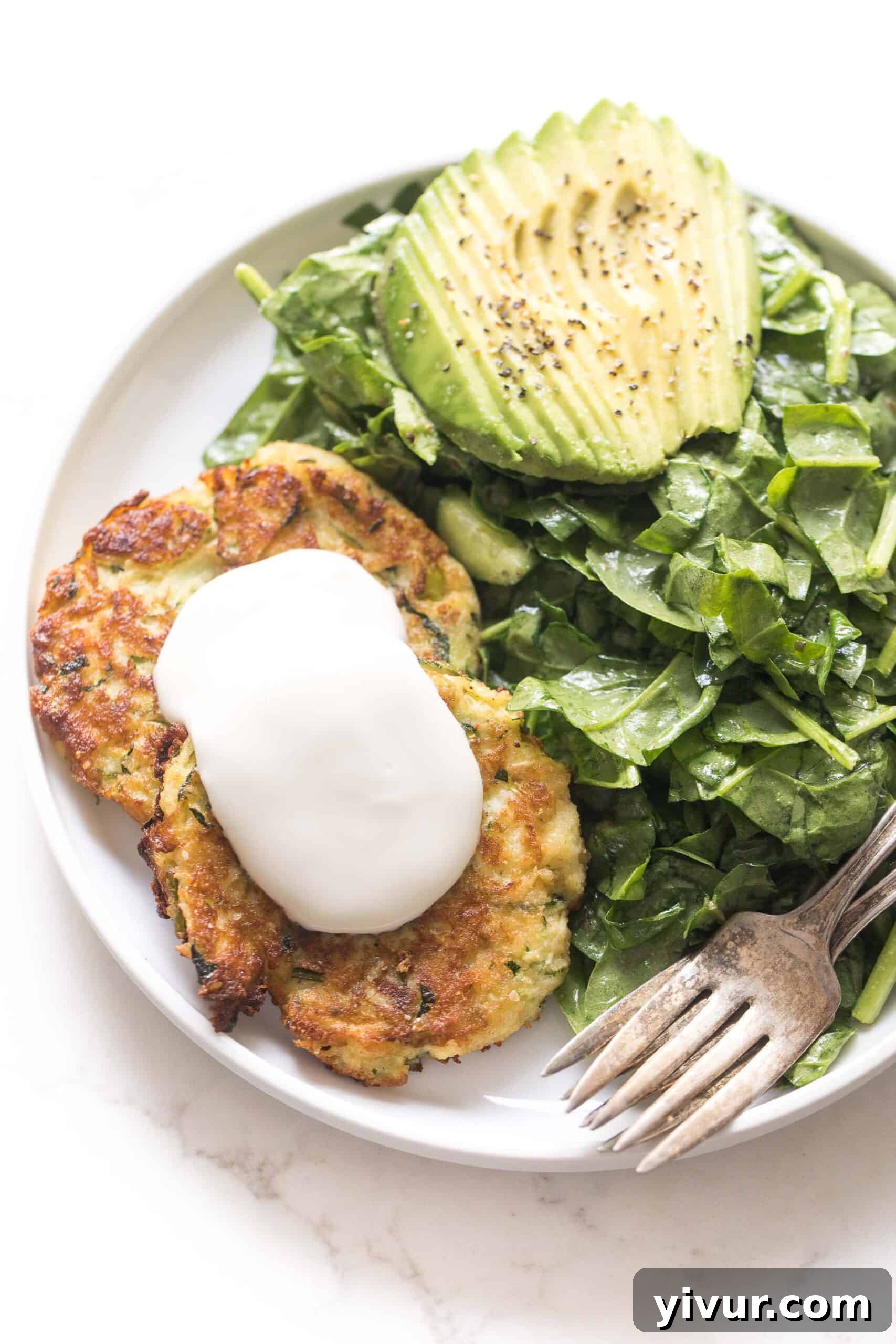 Crispy keto zucchini fritters topped with a dollop of sour cream, served on a white plate with a fresh spinach salad and sliced avocado in the background.