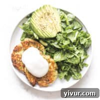 Keto zucchini fritters topped with sour cream on a white plate with a background of spinach salad and avocado.