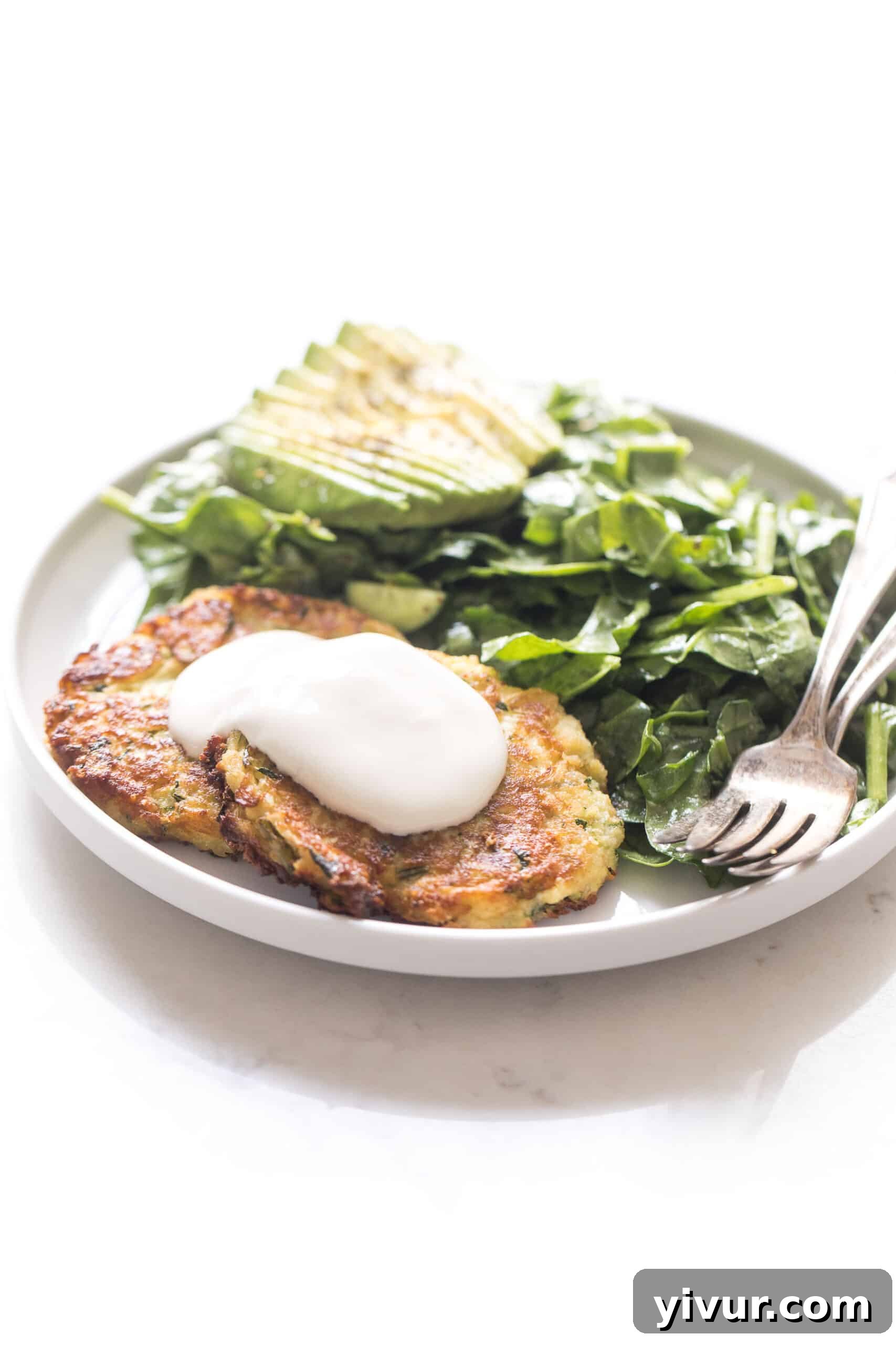 Crispy keto zucchini fritters freshly pan-fried and served with a generous dollop of sour cream, accompanied by a side of vibrant spinach salad and perfectly sliced avocado.