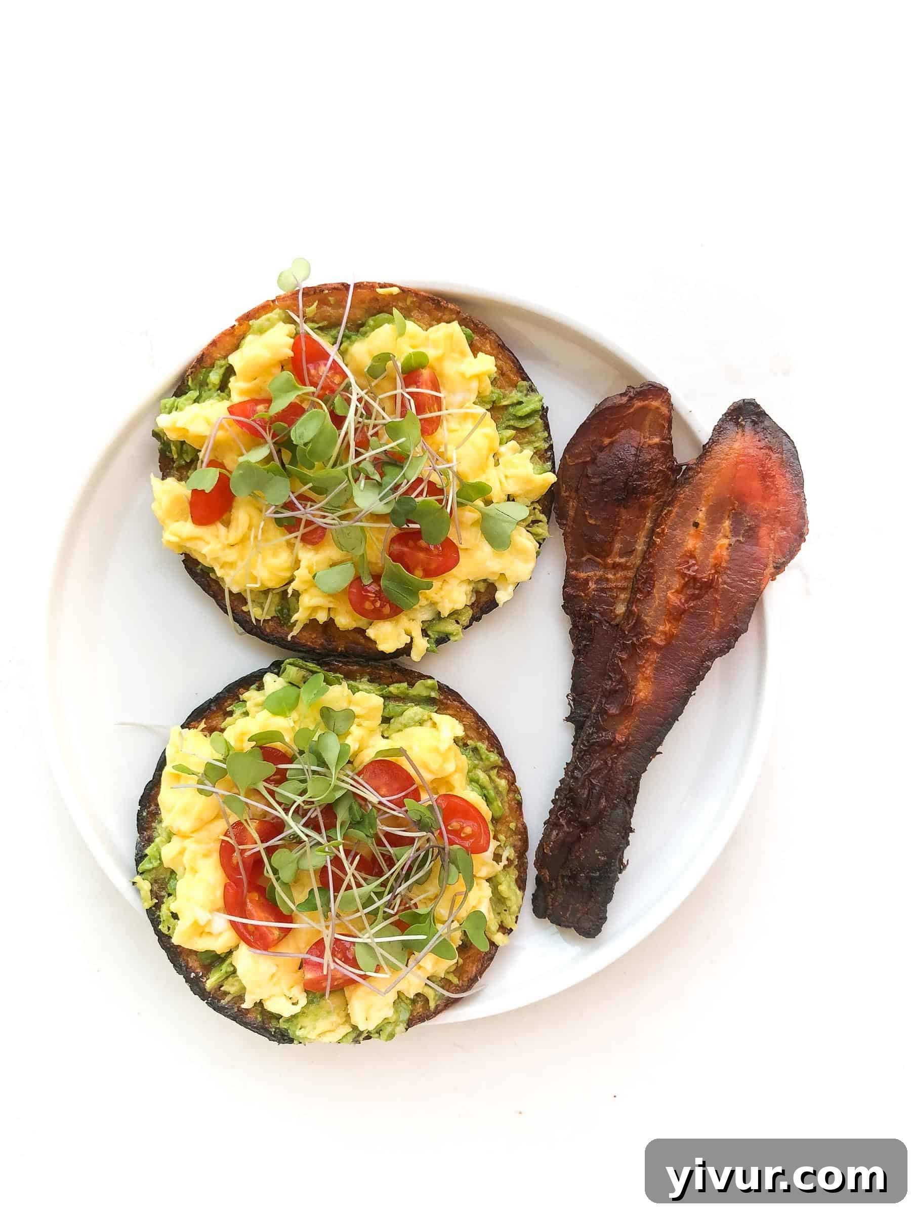 Keto avocado toast topped with scrambled eggs, chopped tomatoes, microgreens, and bacon, served on a white plate.