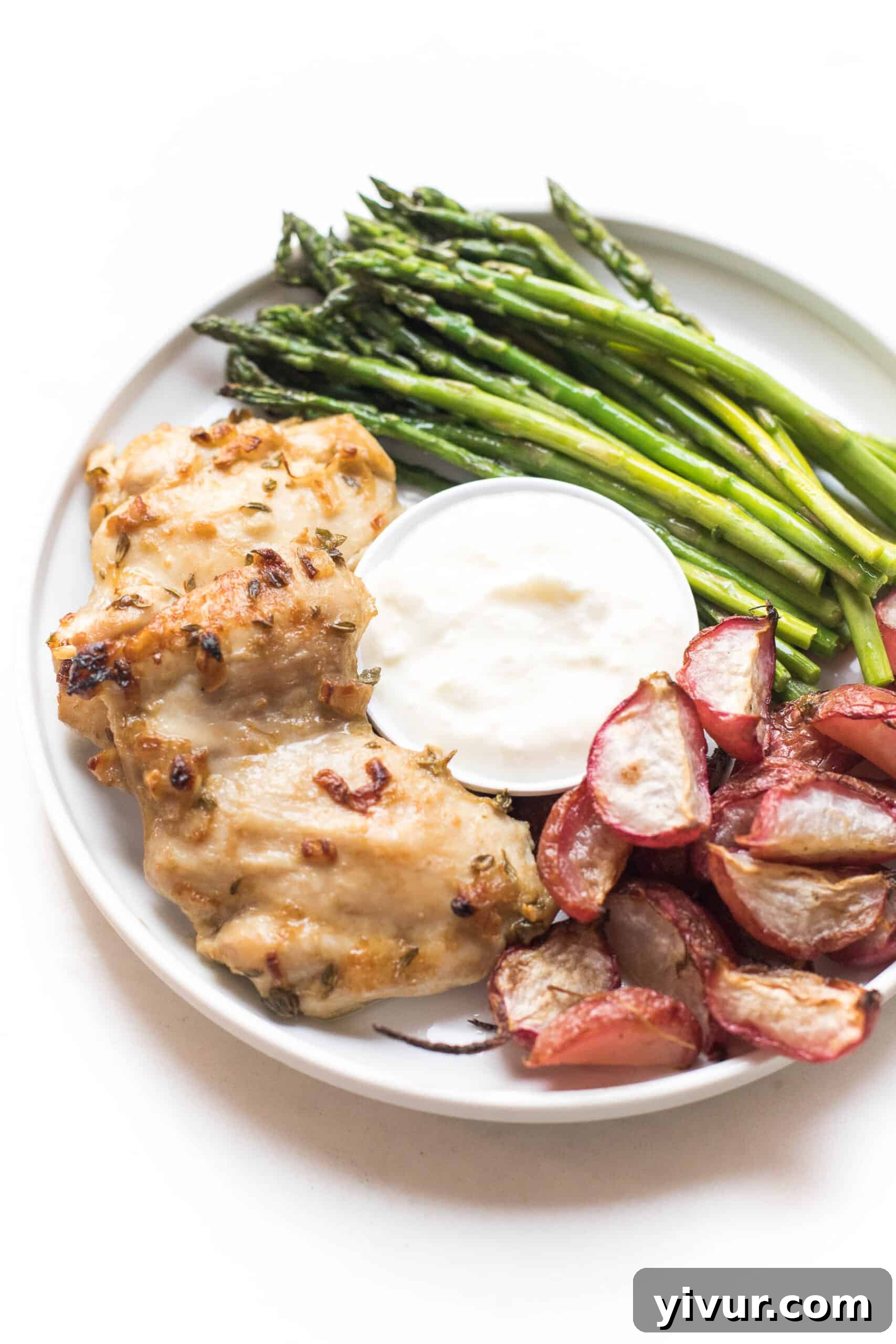 Individual serving of dijon mustard chicken thighs, roasted radishes, and asparagus, elegantly plated with a side of creamy yogurt feta dip on a white dish against a white background.