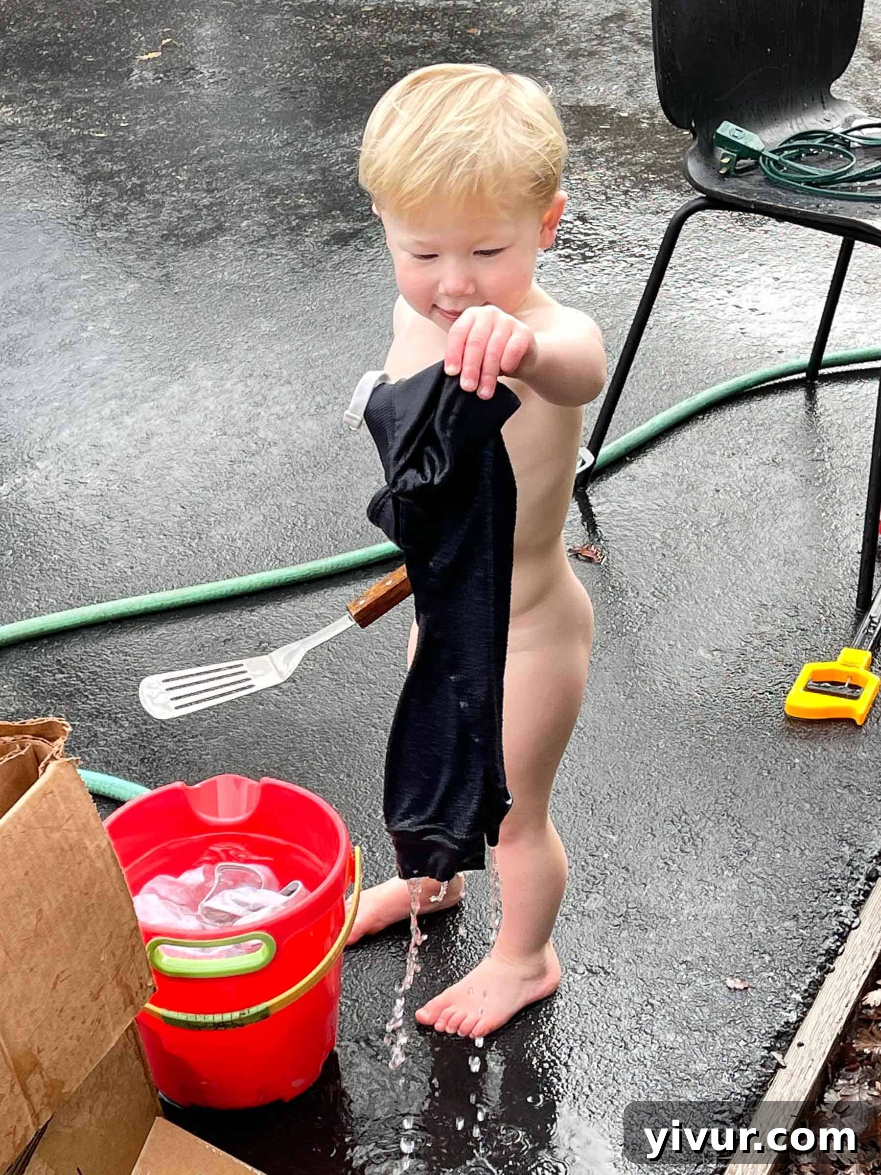 Naked toddler Ryan filling a bucket with water from a hose, pretending to do laundry