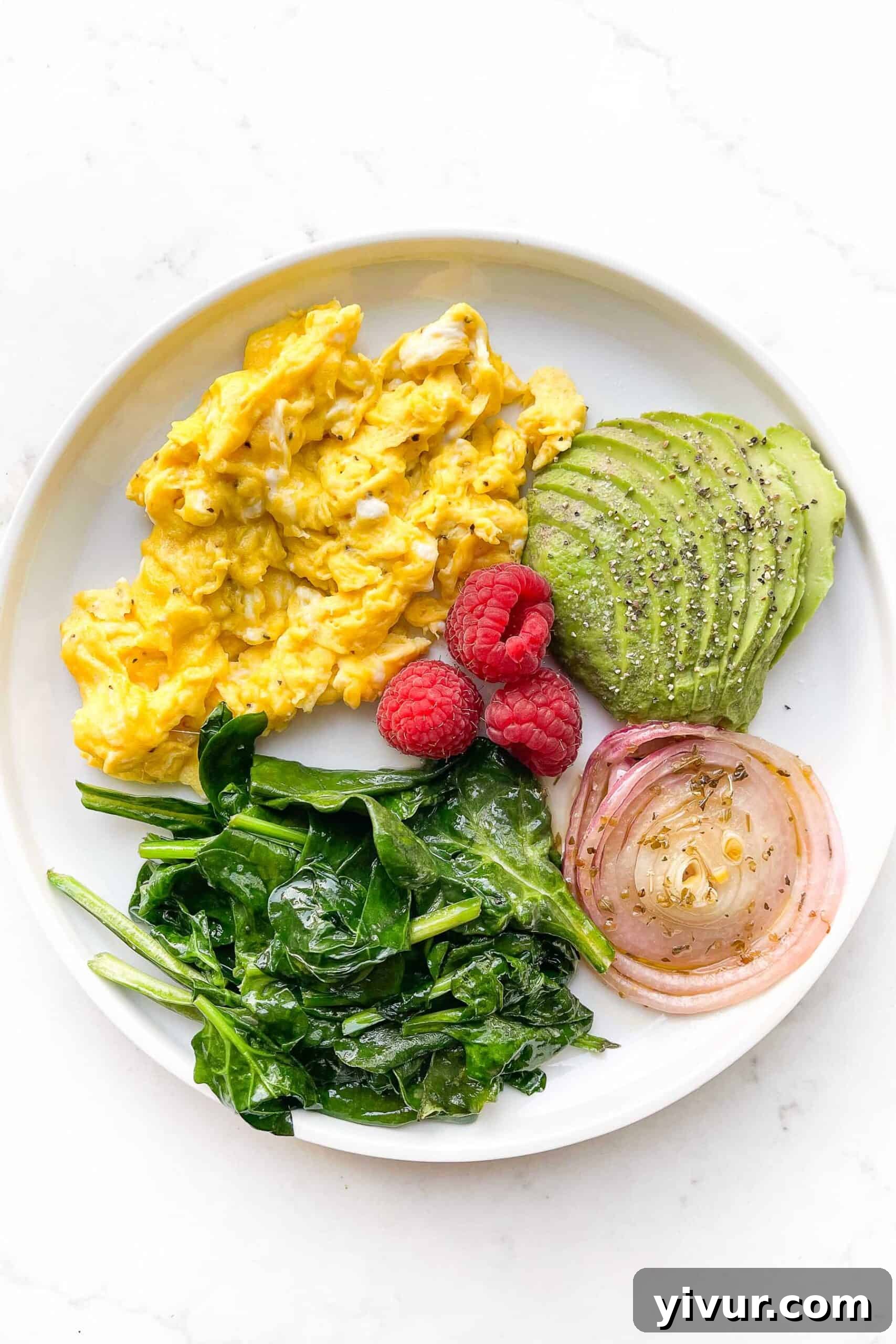 Keto Soft Scrambled Eggs with Spinach and Tangy Marinated Onions 2 A beautifully arranged plate featuring soft scrambled eggs, vibrant sautéed spinach, creamy avocado slices, bright red marinated onions, and a few fresh raspberries, set against a clean white background.
