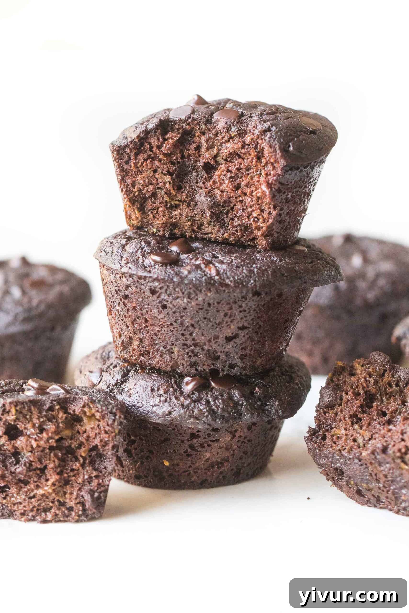 A stack of perfectly baked keto chocolate zucchini muffins, with one showing a delightful bite taken out, set against a clean white background, tempting to eat.