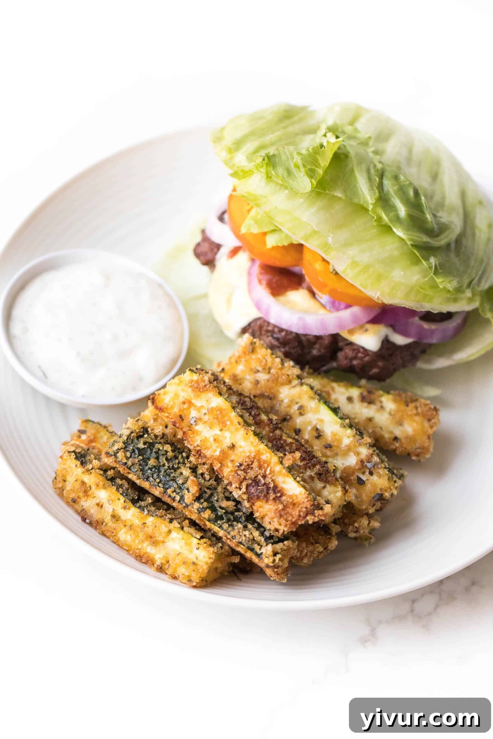 Crispy keto zucchini fries served on a white plate alongside a juicy burger and a small bowl of creamy ranch dressing, highlighting a delicious low-carb meal option.