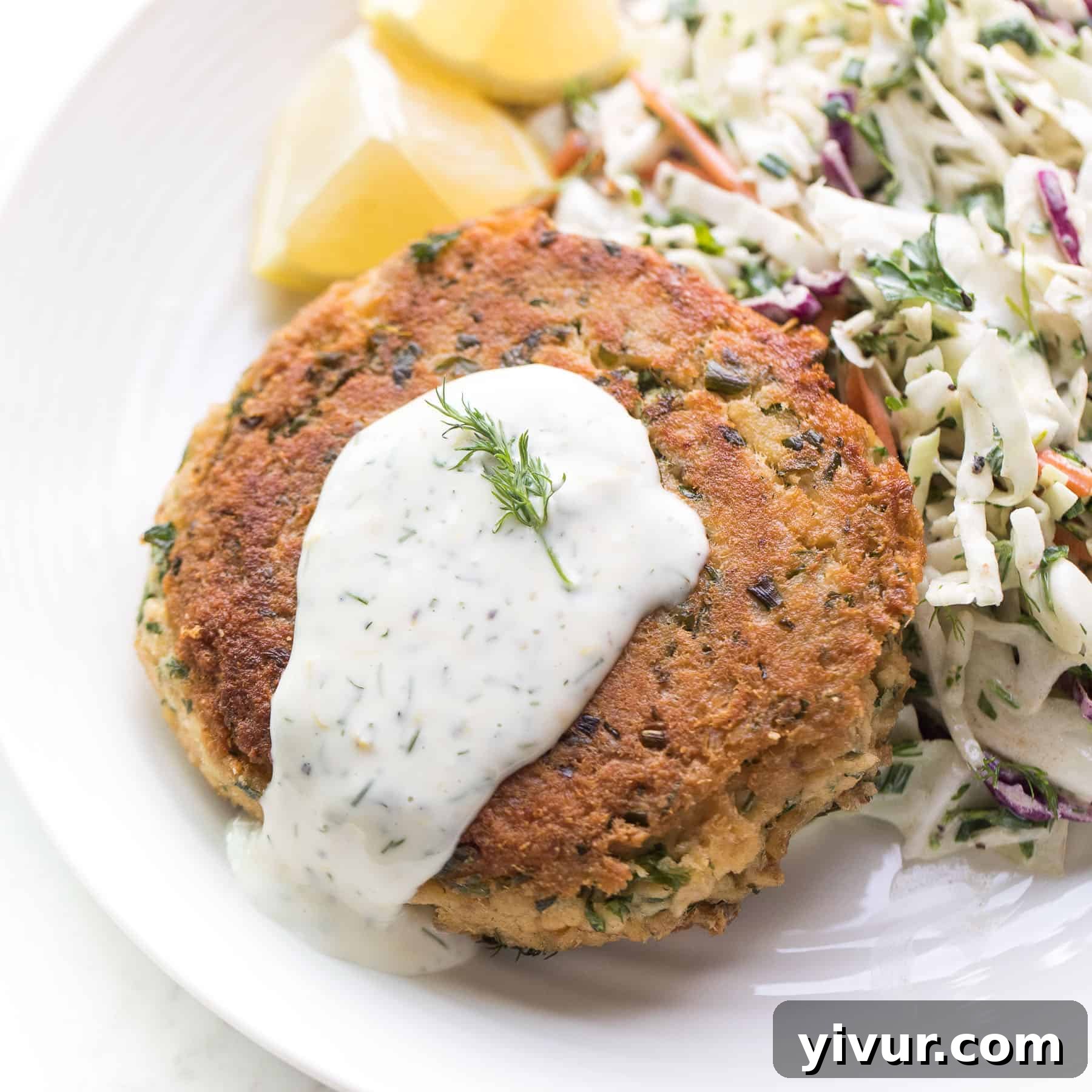 salmon patty with white sauce on a white plate with lemon and cabbage salad