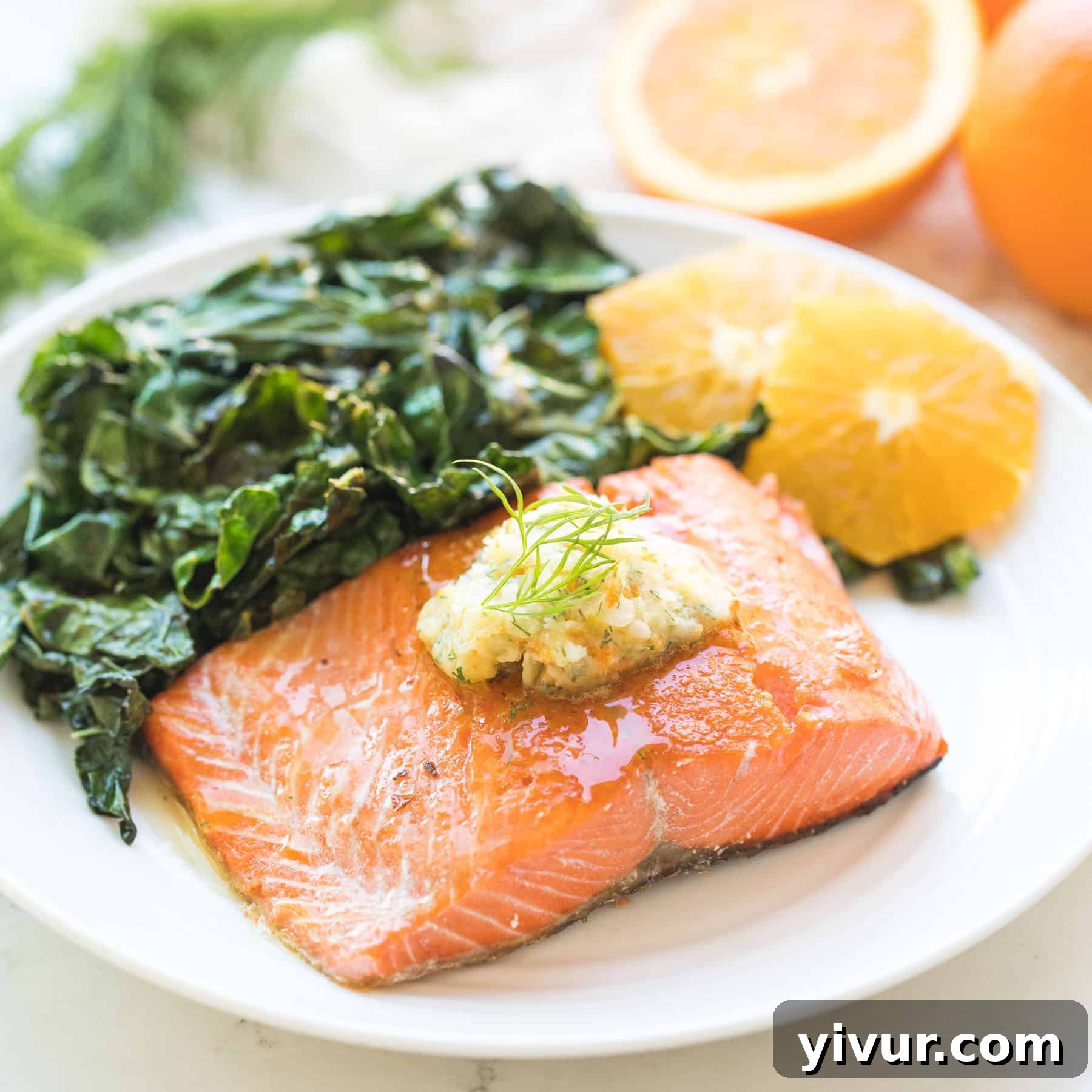 salmon on a white plate with butter on top next to peeled oranges and green kale salad