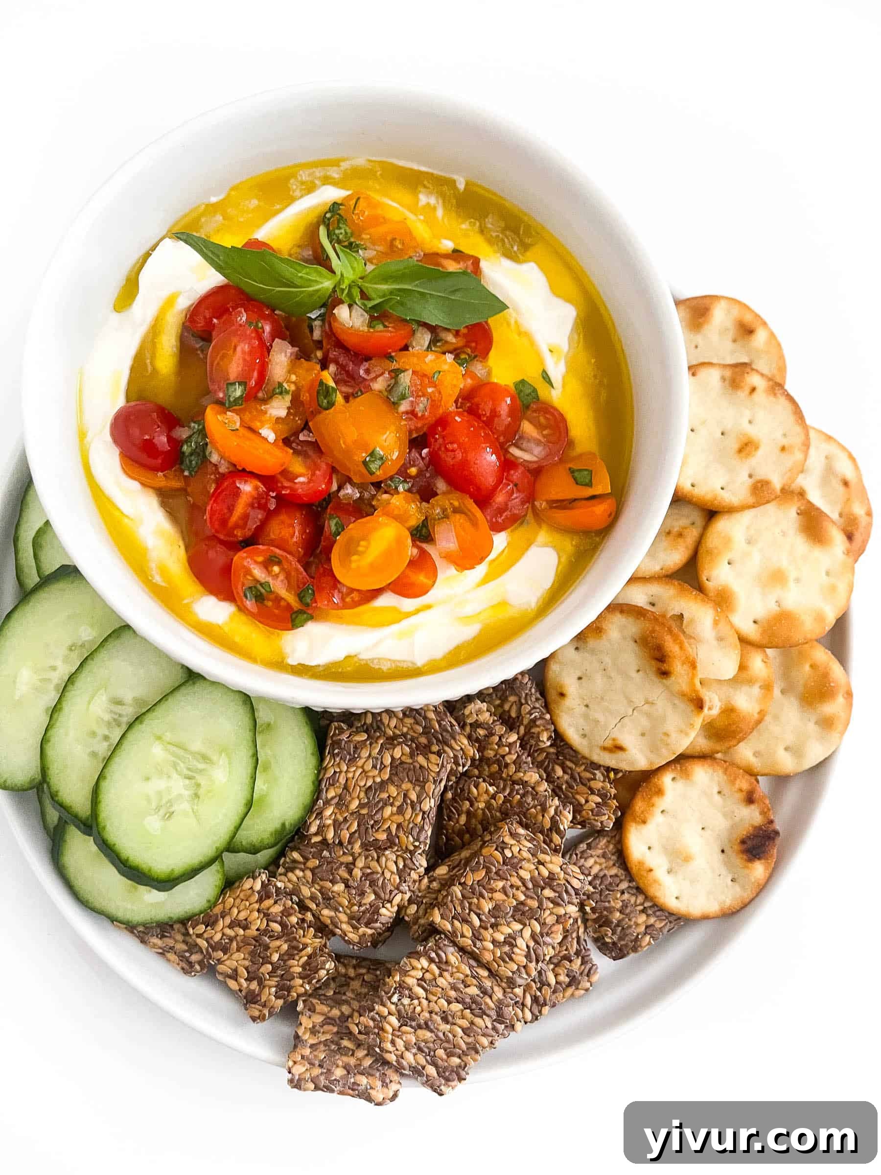 Whipped ricotta dip in a white bowl topped with grape tomatoes, basil and olive oil with cucumbers and crackers