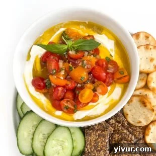 Whipped ricotta dip in a white bowl topped with grape tomatoes, basil and olive oil with cucumbers and crackers
