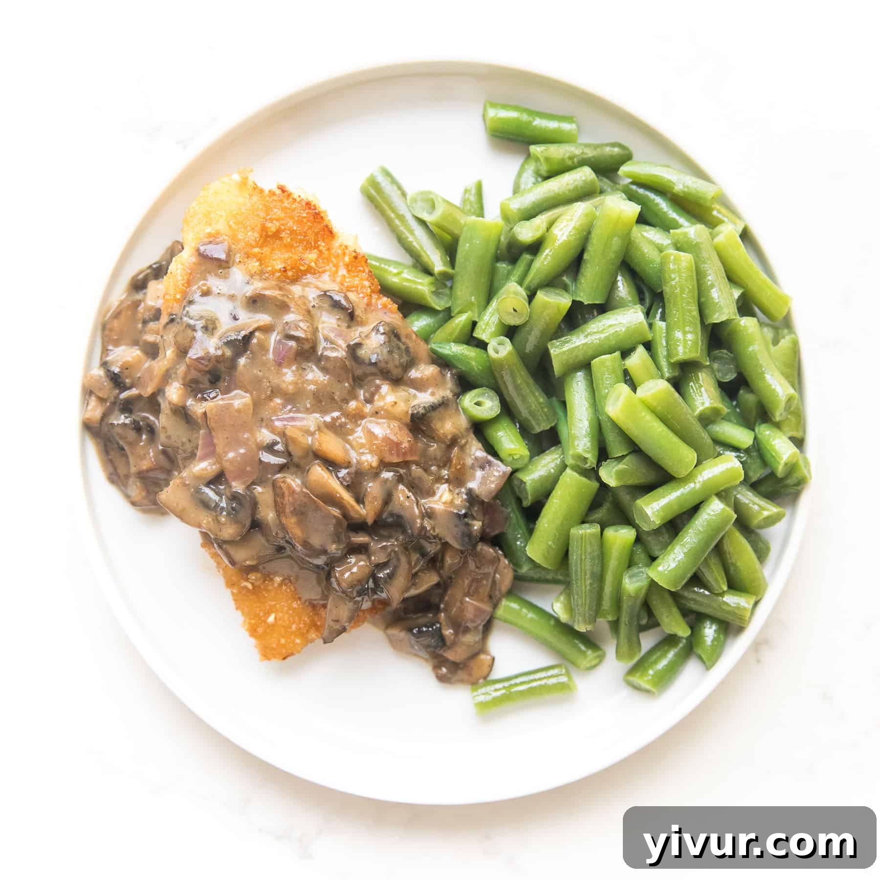 Golden breaded chicken with rich mushroom gravy on a white plate, served alongside green beans.