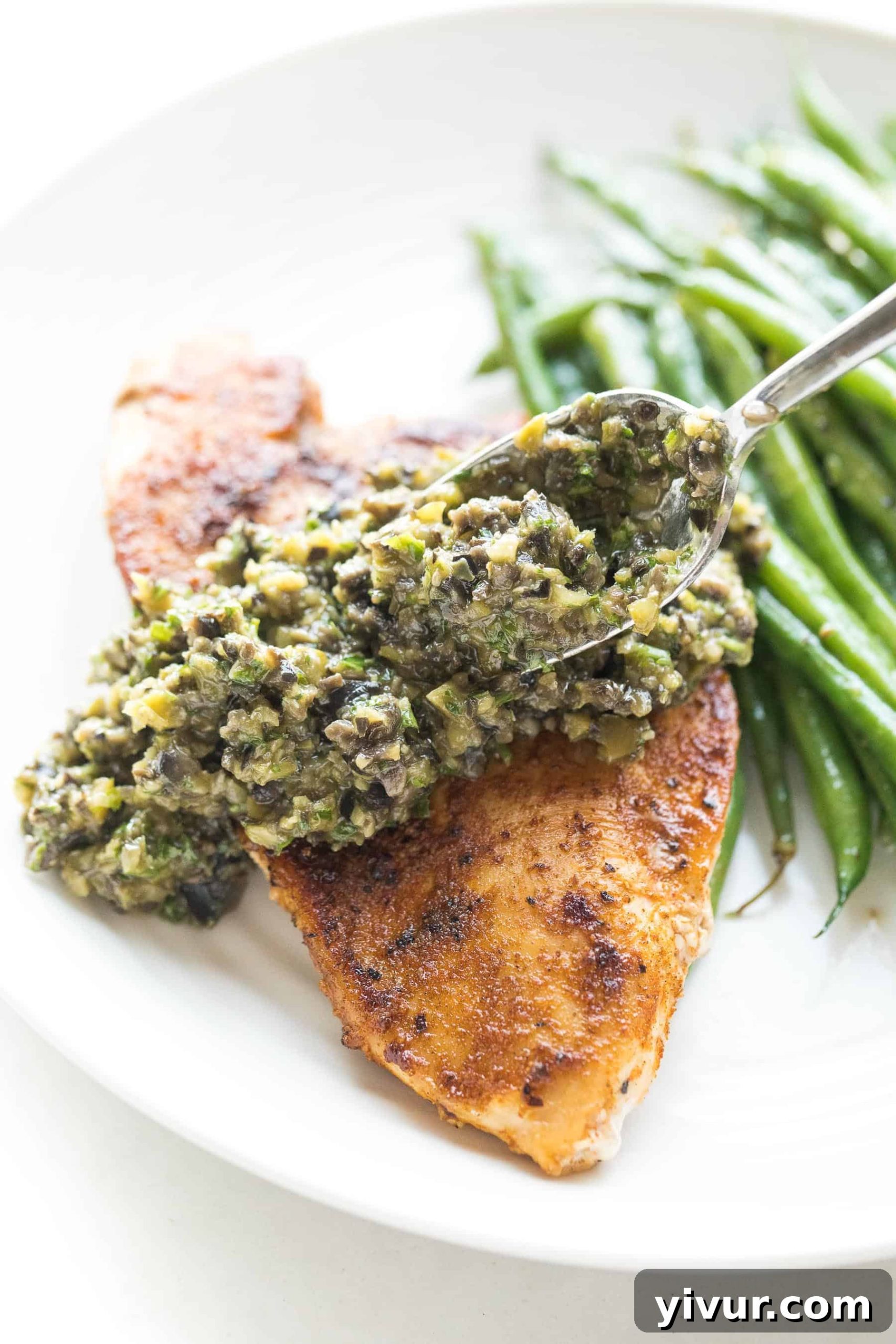 A spoon putting olive tapenade on top of succulent chicken with green beans in the background. Paleo + Whole30 Chicken with Olive Tapenade - savory seasoned chicken topped with a bright and fresh olive tapenade. A healthy and delicious 30 minute chicken dinner recipe! Gluten free, grain free, dairy free, sugar free, clean eating, real food. #paleo #whole30 #chicken