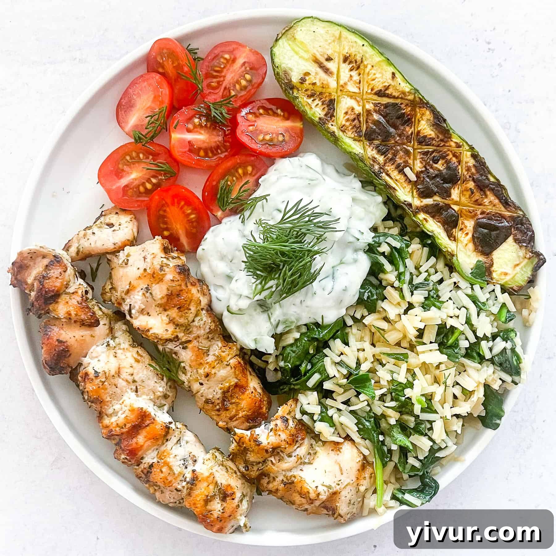 Keto grilled chicken kabobs on a white plate with fresh veggies and a dollop of tzatziki sauce on a white background.