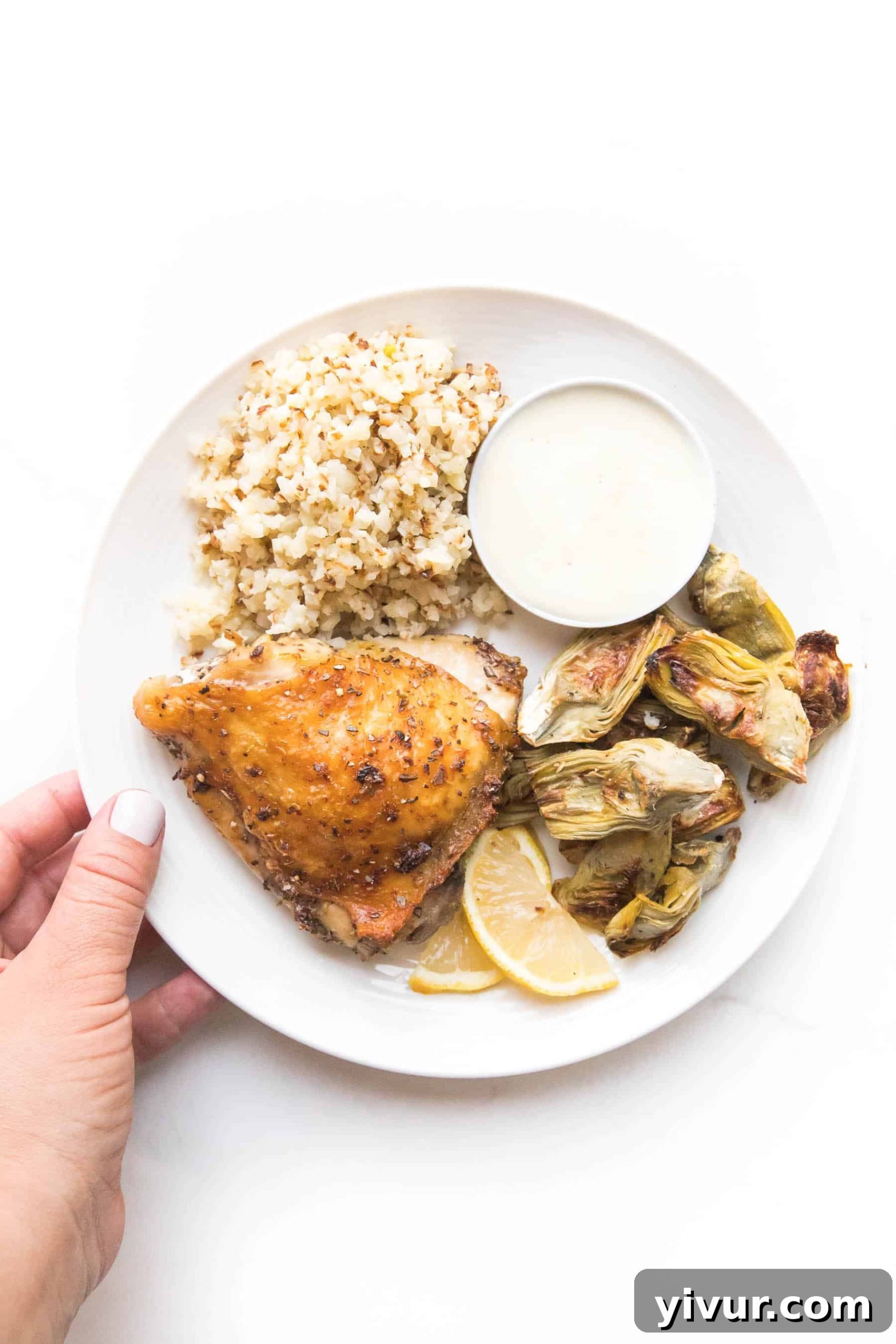 A hand holding a white plate with a chicken thigh, roasted artichoke hearts, and cauliflower rice, showcasing a Keto Sheet Pan Greek Chicken meal.