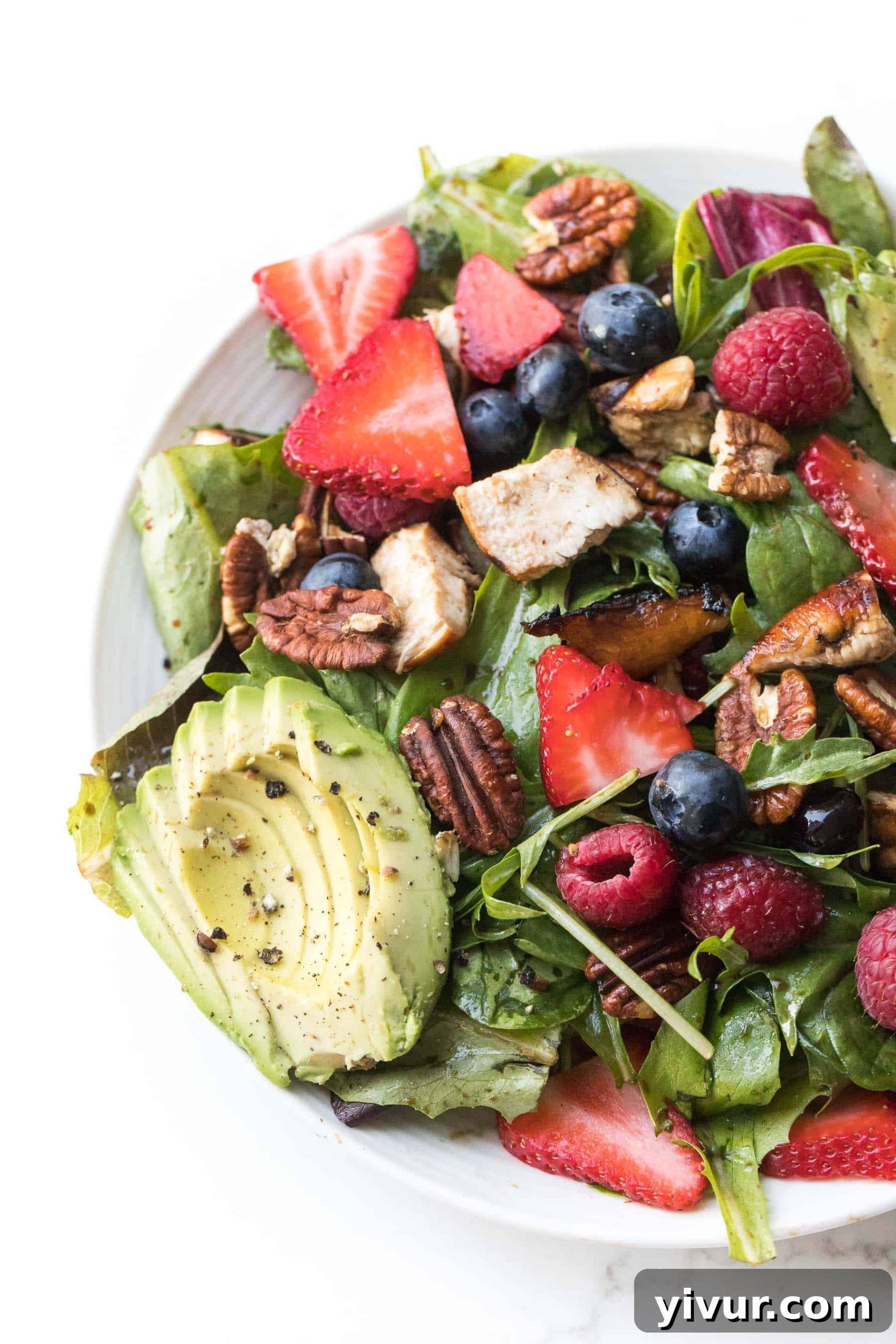 A vibrant salad with strawberries, blueberries, raspberries, grilled chicken, creamy avocado, and crunchy pecans on a white plate and background.