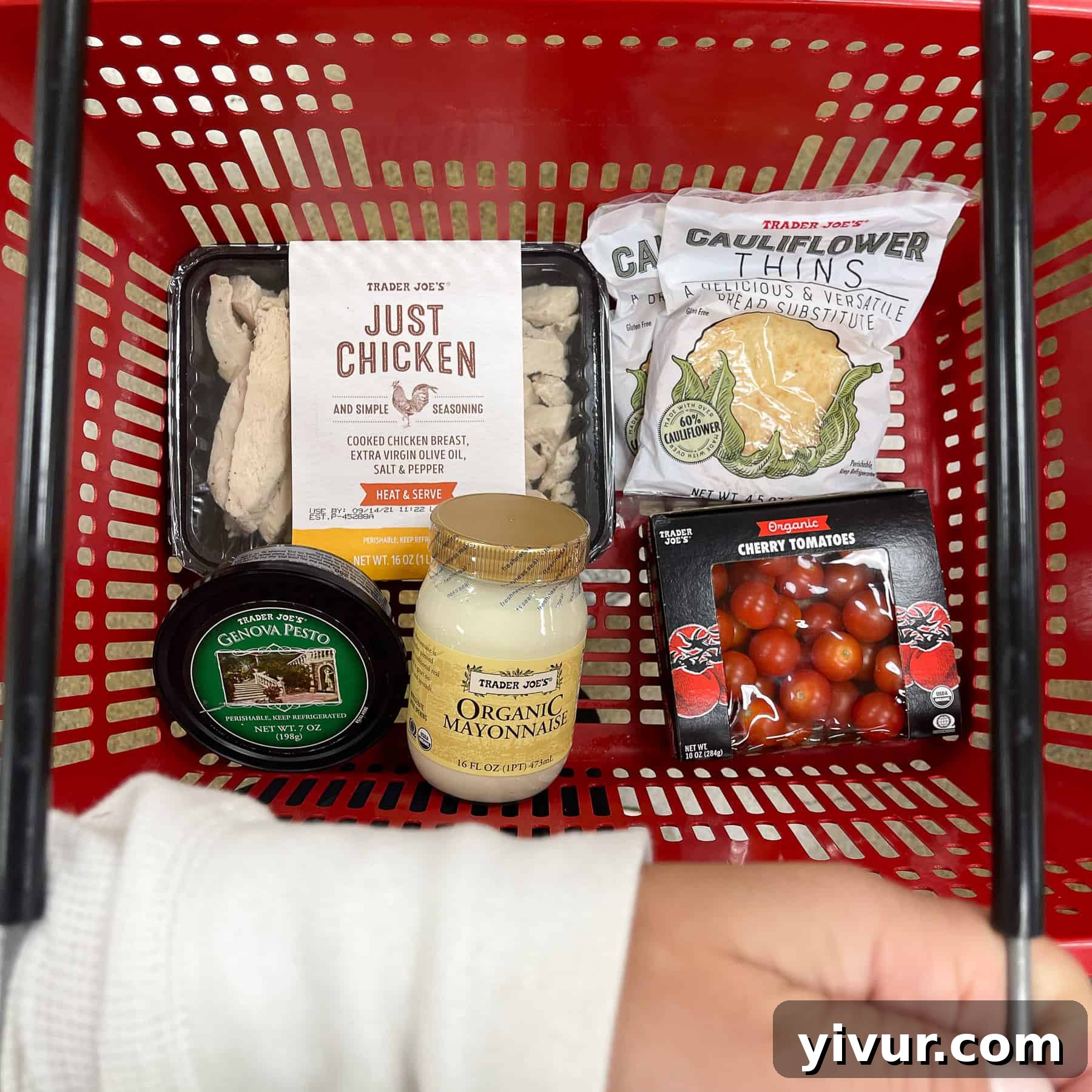 A vibrant red Trader Joe's basket filled with the five essential ingredients for Keto Pesto Chicken Salad: cooked chicken, mayonnaise, cherry tomatoes, pesto, and cauliflower thins, ready for preparation.