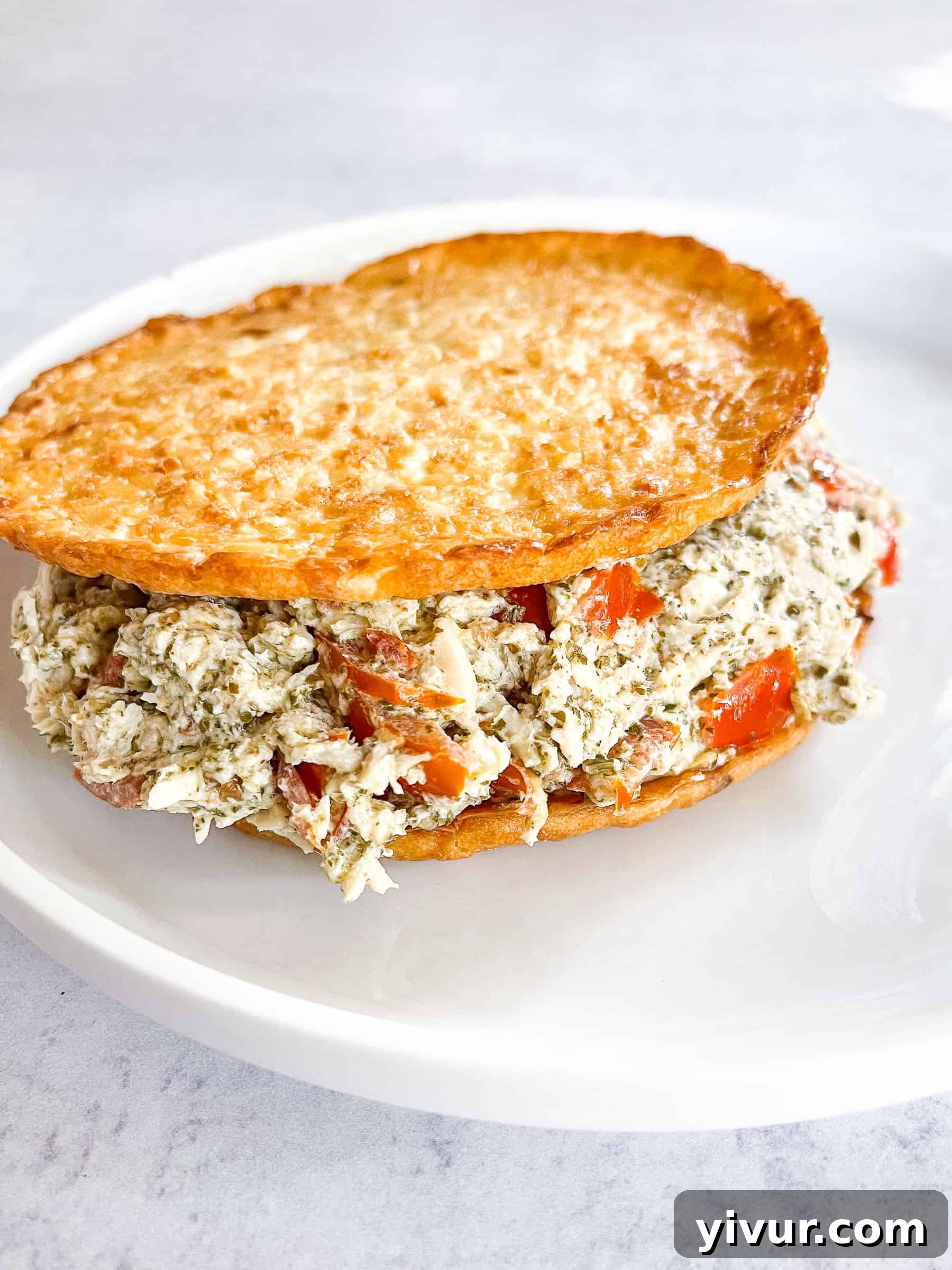 The finished Keto Pesto Chicken Salad generously spread on two cauliflower thins, served elegantly on a white plate against a minimalist white background, ready to be enjoyed as a low-carb meal.