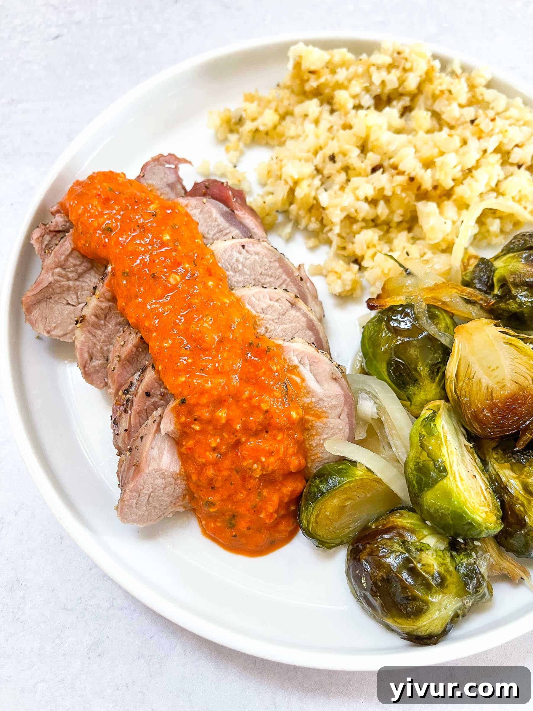 Keto Sheet Pan Pork Tenderloin with Romesco on a white plate with Brussel sprouts and cauliflower rice, set against a clean white background.