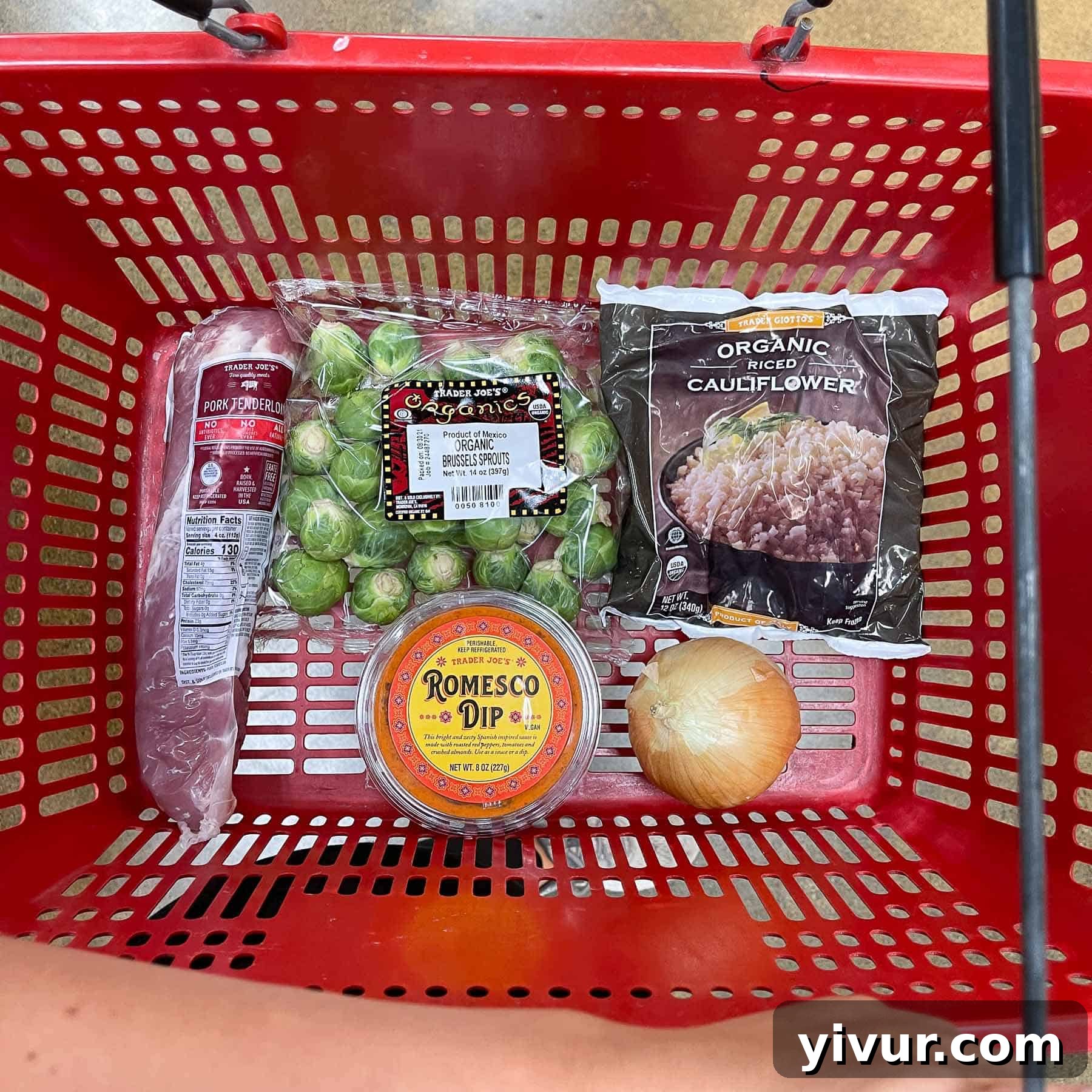 A red Trader Joe's basket overflowing with fresh ingredients for Keto Sheet Pan Pork Tenderloin with Romesco, including pork tenderloin, Brussels sprouts, onions, cauliflower rice, and a jar of romesco dip.