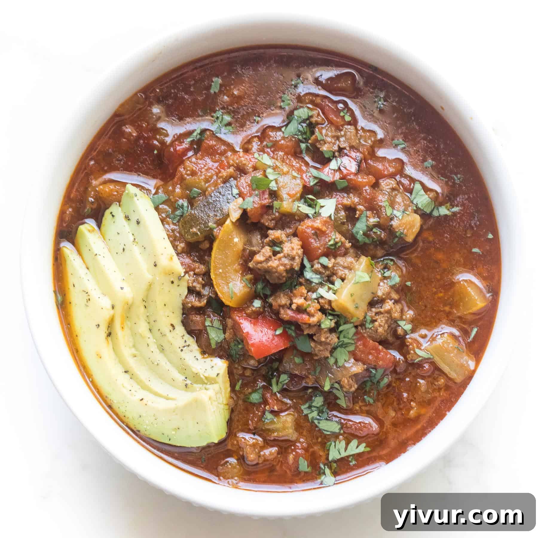 Rich keto chili in a white bowl, garnished with vibrant green herbs and fresh avocado slices, against a clean white background.