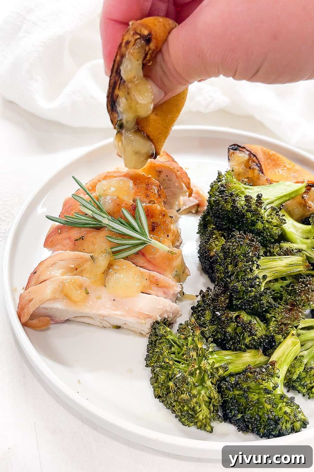 a hand squeezing a grilled lemon on top of sliced flattened chicken on a white plate next to a pile of roasted broccoli