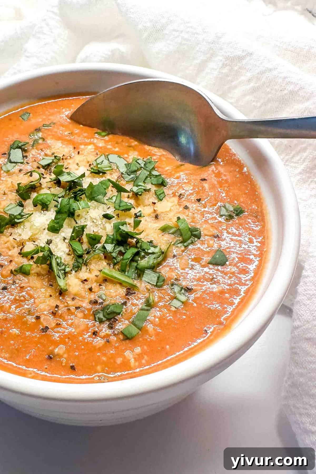 A spoon scooping creamy tomato basil soup out of a white bowl, showing its thick and luxurious texture
